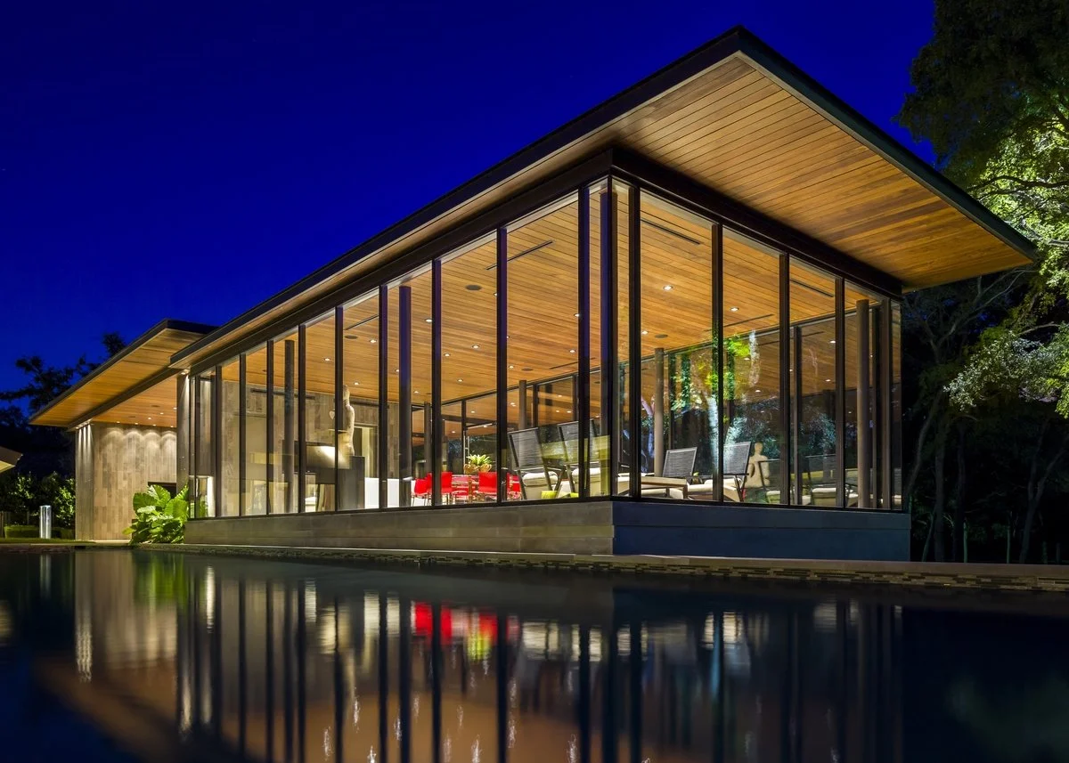 Modern house with glass walls and wooden ceiling, illuminated at night, with a reflection in the pool in the foreground, surrounded by trees.