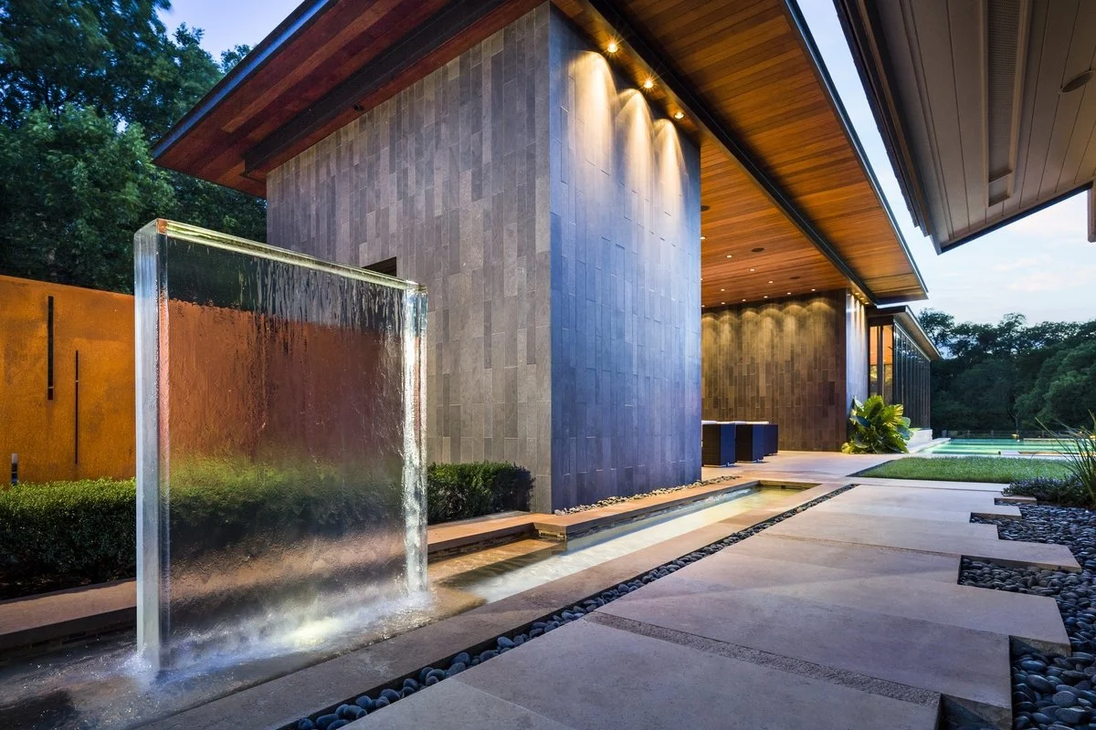 Modern pool house exterior with acrylic water feature, waterfall, landscaping, and outdoor lighting at dusk.