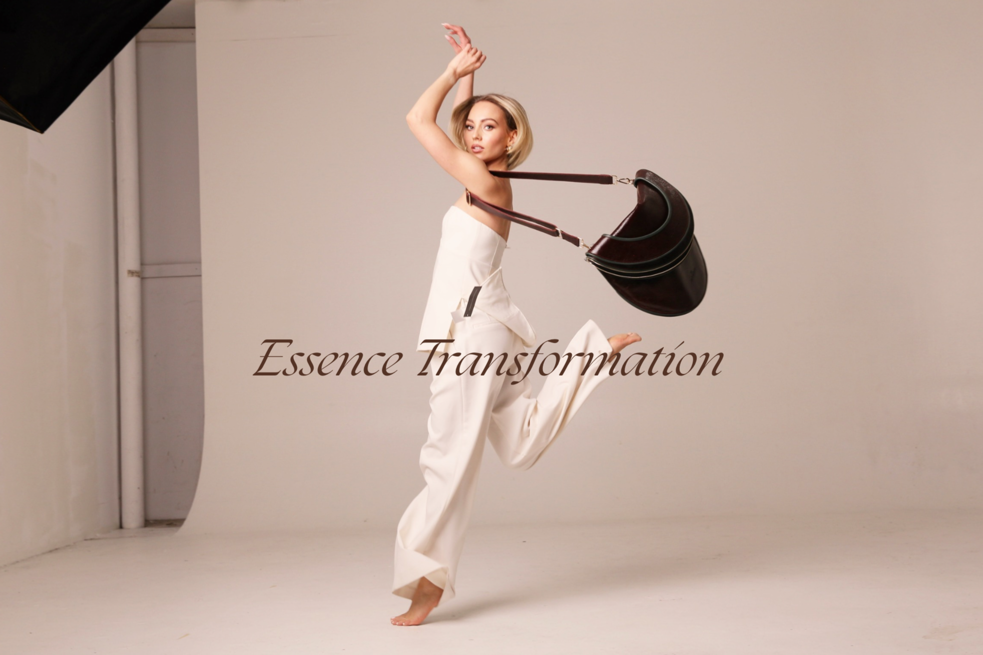 A woman in a white dress is jumping in a photo studio with a gray background, carrying a large brown handbag. The text 'Essence Transformation' is overlaid on the image.