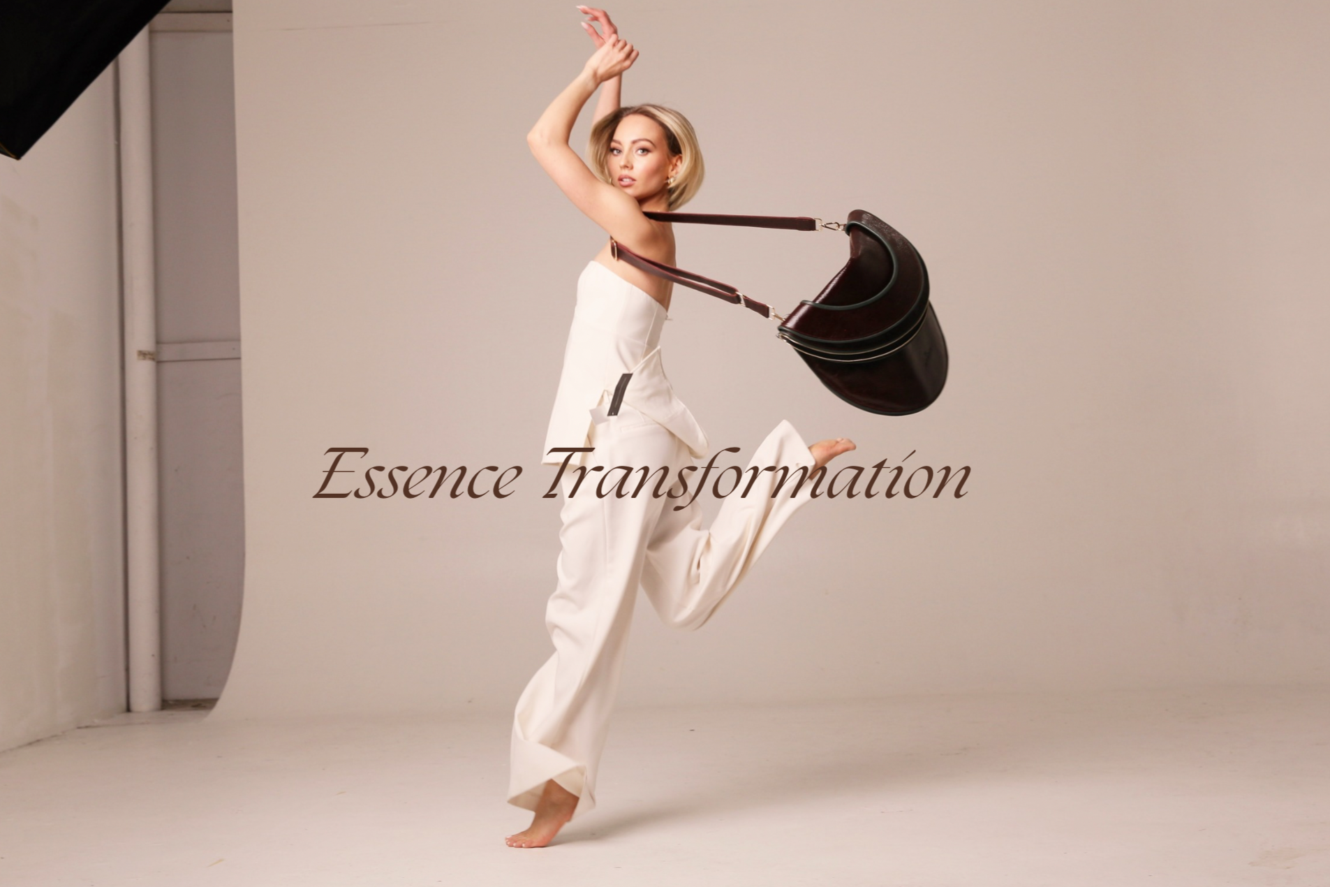 A woman in a white dress jumps in a photo studio, with a large brown handbag slung over her shoulder, and the words 'Essence Transformation' written across her midsection.
