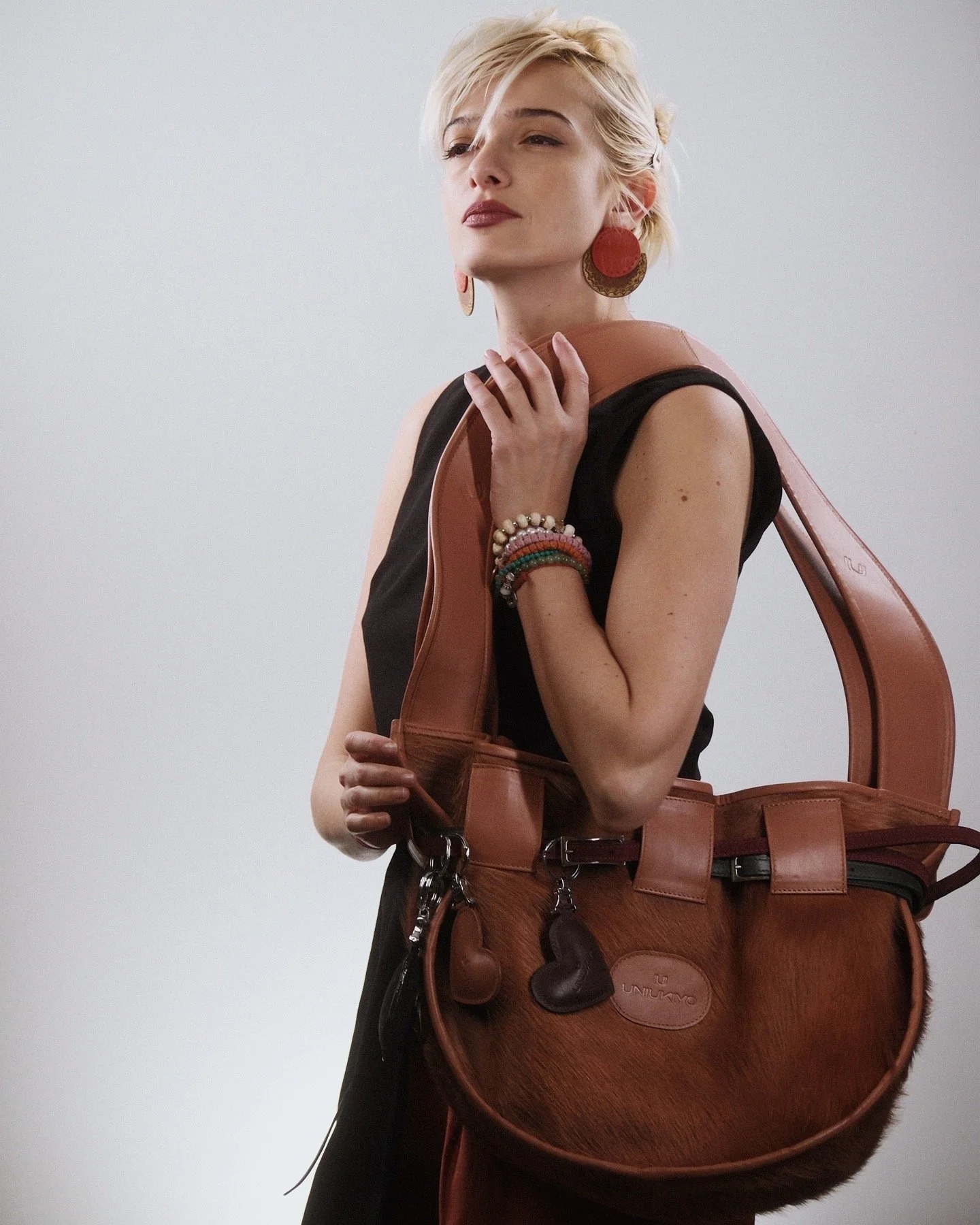 A woman with short blonde hair and wearing large red earrings, a black sleeveless dress, and colorful bracelets on her wrist, holds a large brown leather handbag with a fur texture, a heart-shaped keychain, and other small accessories, standing against a plain light background.