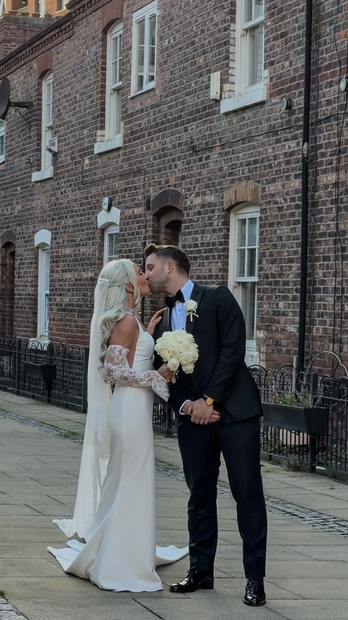 A bride and groom sharing a kiss outdoors on a city sidewalk, with brick buildings in the background. The bride is wearing a white wedding dress with lace sleeves and holding a bouquet of white roses. The groom is dressed in a black tuxedo with a bow tie and boutonniere.