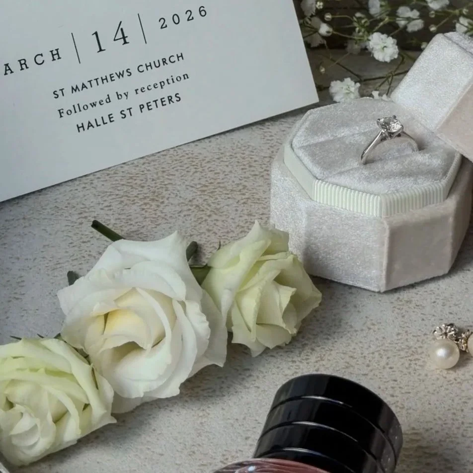 A wedding invitation for March 14, 2026, at St. Matthews Church with reception at Halle St. Peters, along with white roses, a ring in a white velvet box, and pearl earrings displayed on a textured surface.