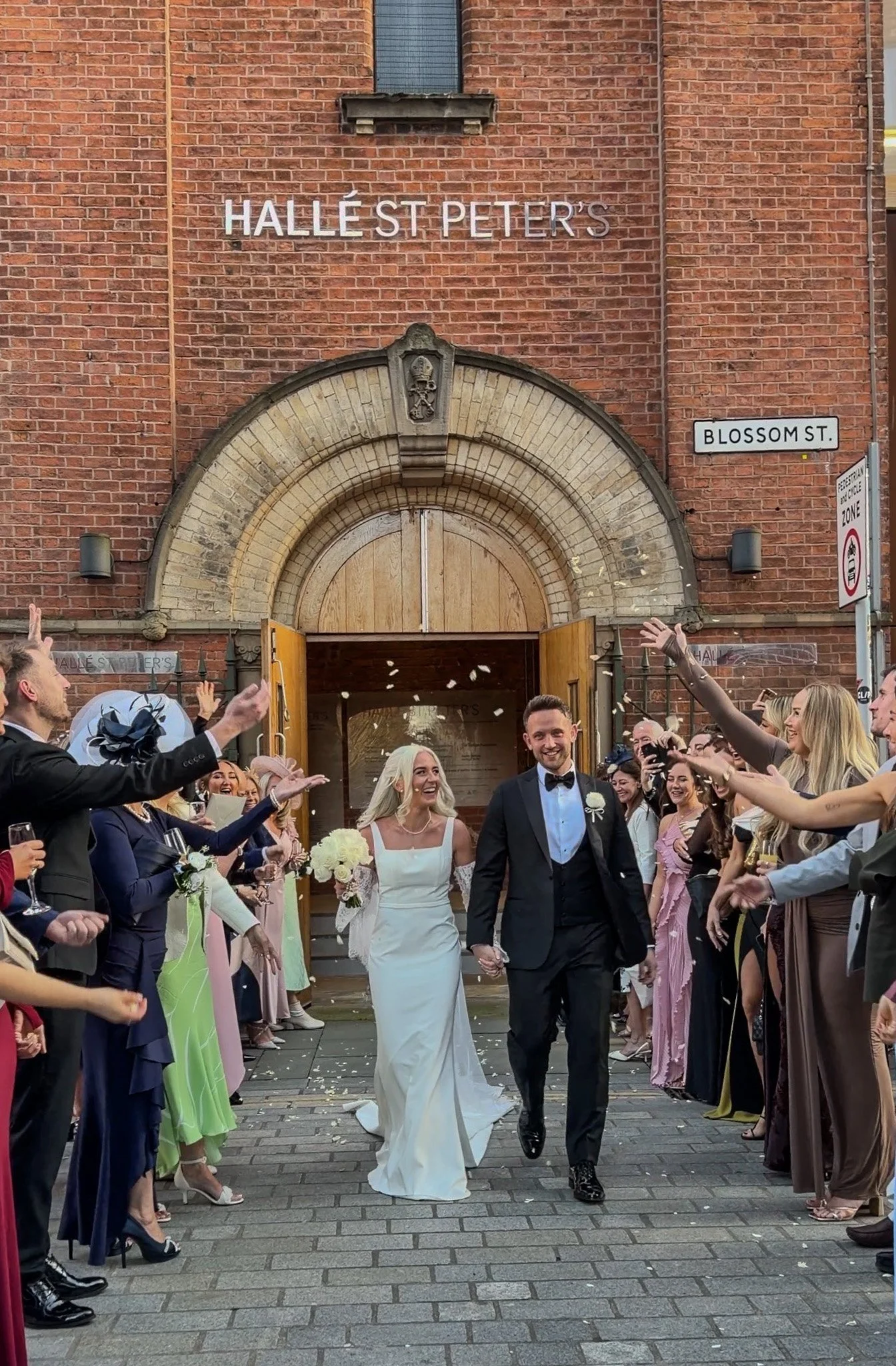 Bride and groom walking out of a church, smiling and holding hands, surrounded by guests throwing confetti on a city street.