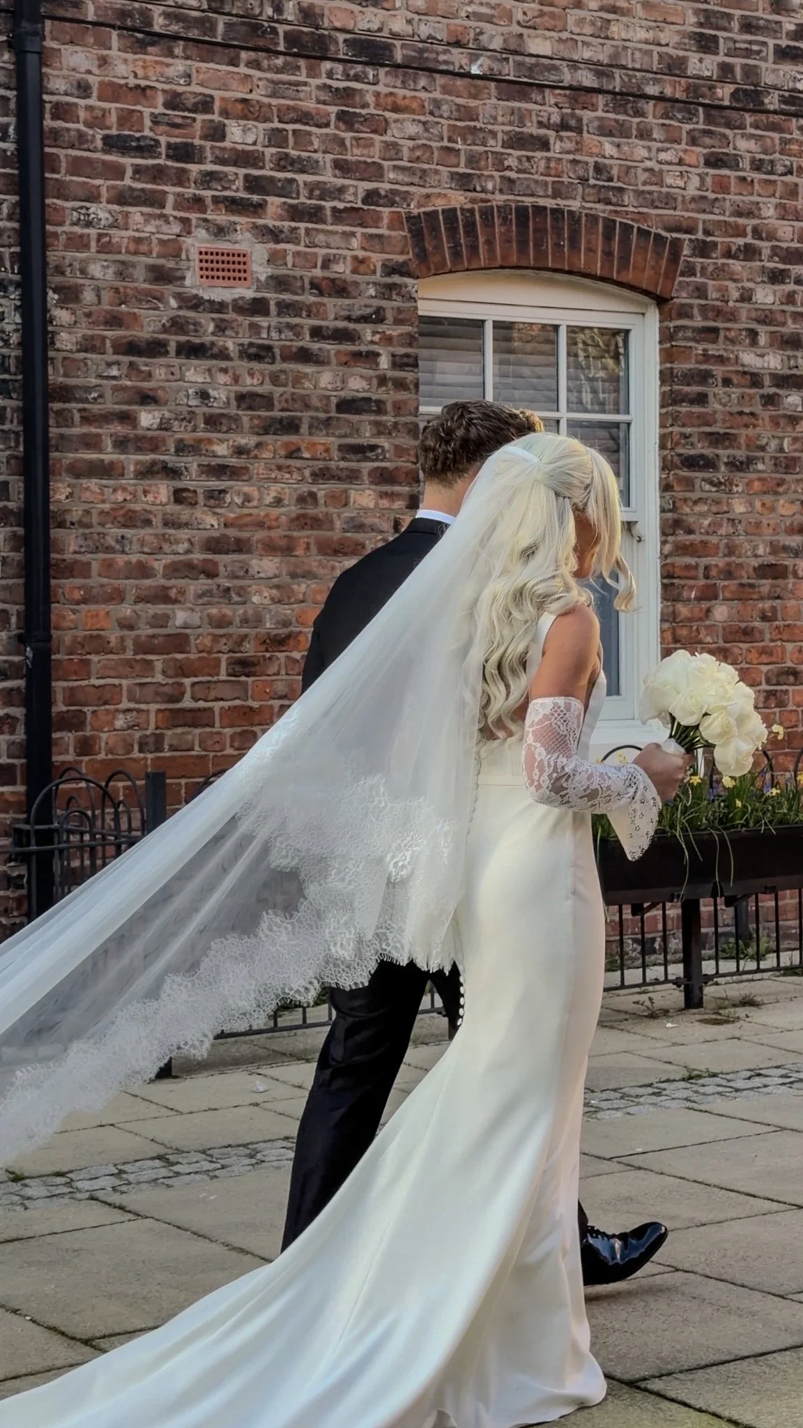 Bride in a white wedding gown with lace sleeves and a long veil holding a bouquet of white roses, next to groom in a black suit outside in front of a brick building.