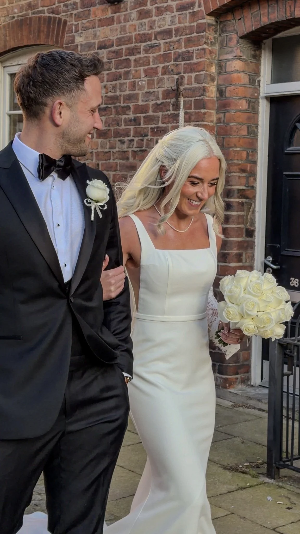 A bride with long platinum blonde hair smiling and holding a bouquet of white roses, arm linked with a groom wearing a black tuxedo, light blue shirt, and black bowtie, standing in front of a brick building.