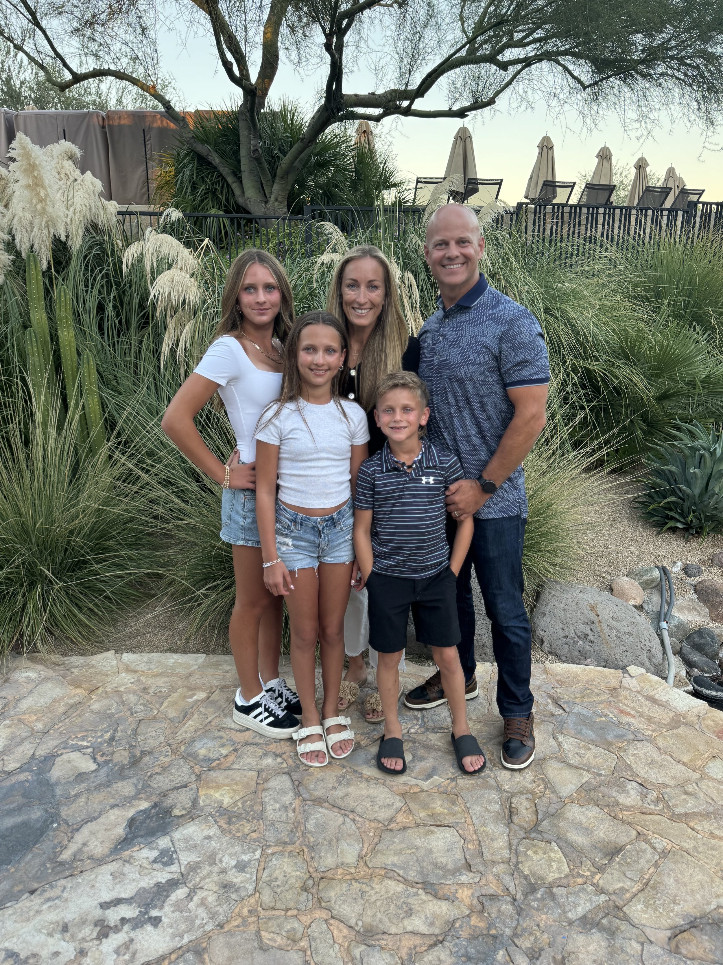 A family of six standing on a stone patio outdoors, surrounded by tall grasses, trees, and an outdoor patio area with umbrellas in the background.