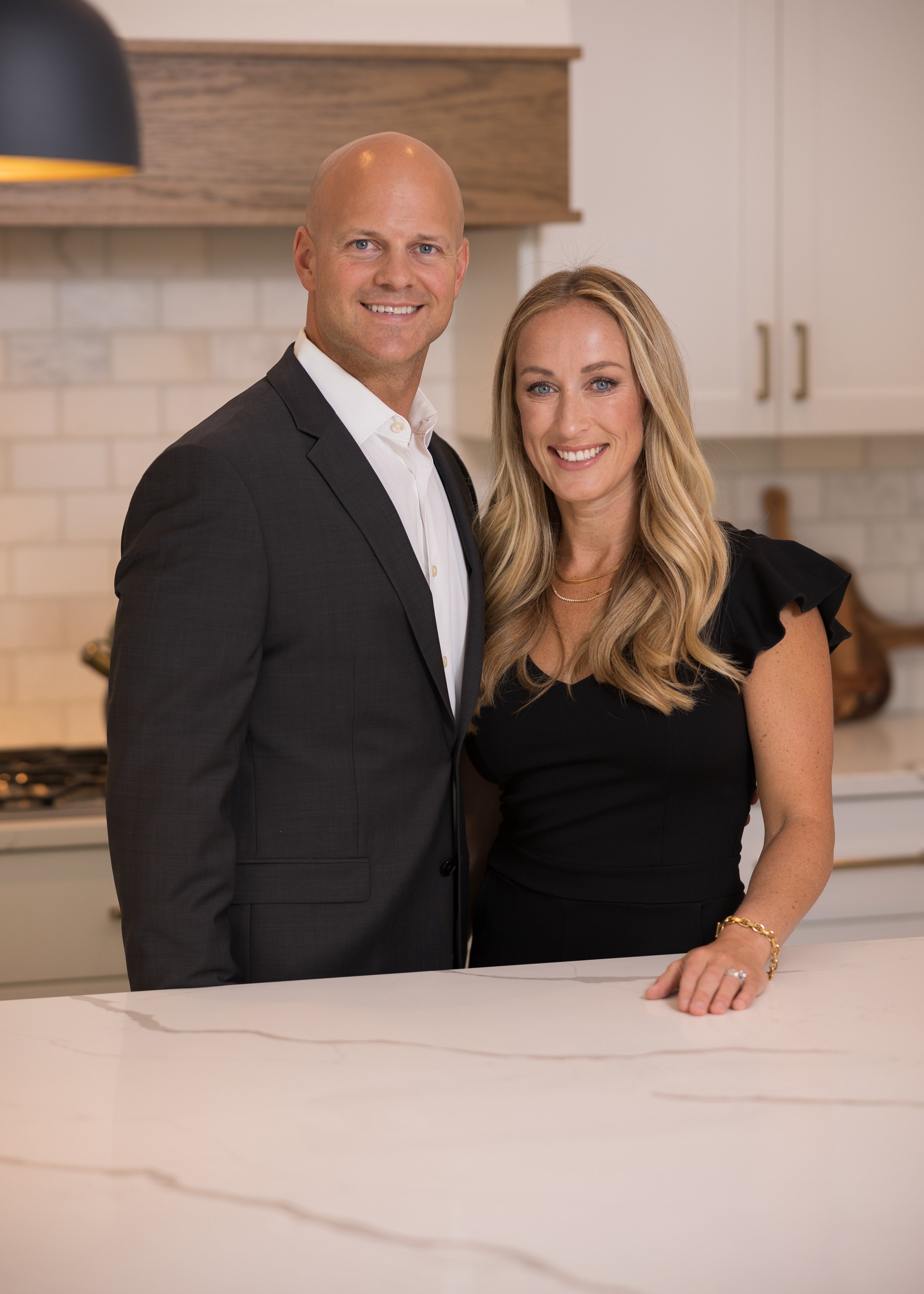 A man in a dark suit and a woman in a black dress standing together in a kitchen.