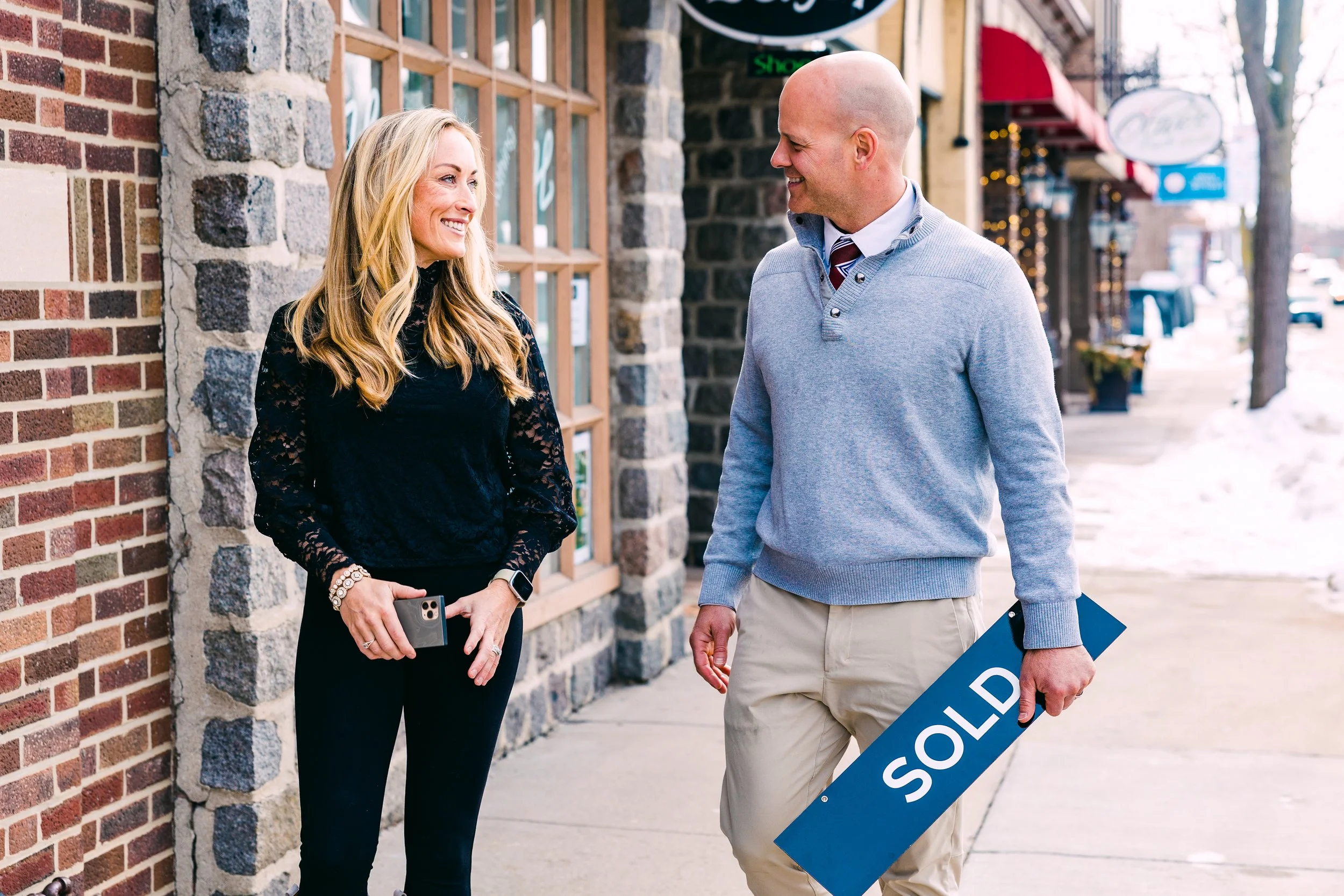 A man wearing a gray sweater and khaki pants holding a blue 'SOLD' sign, smiling and talking to a woman dressed in black, standing outside a storefront with a brick and stone wall.