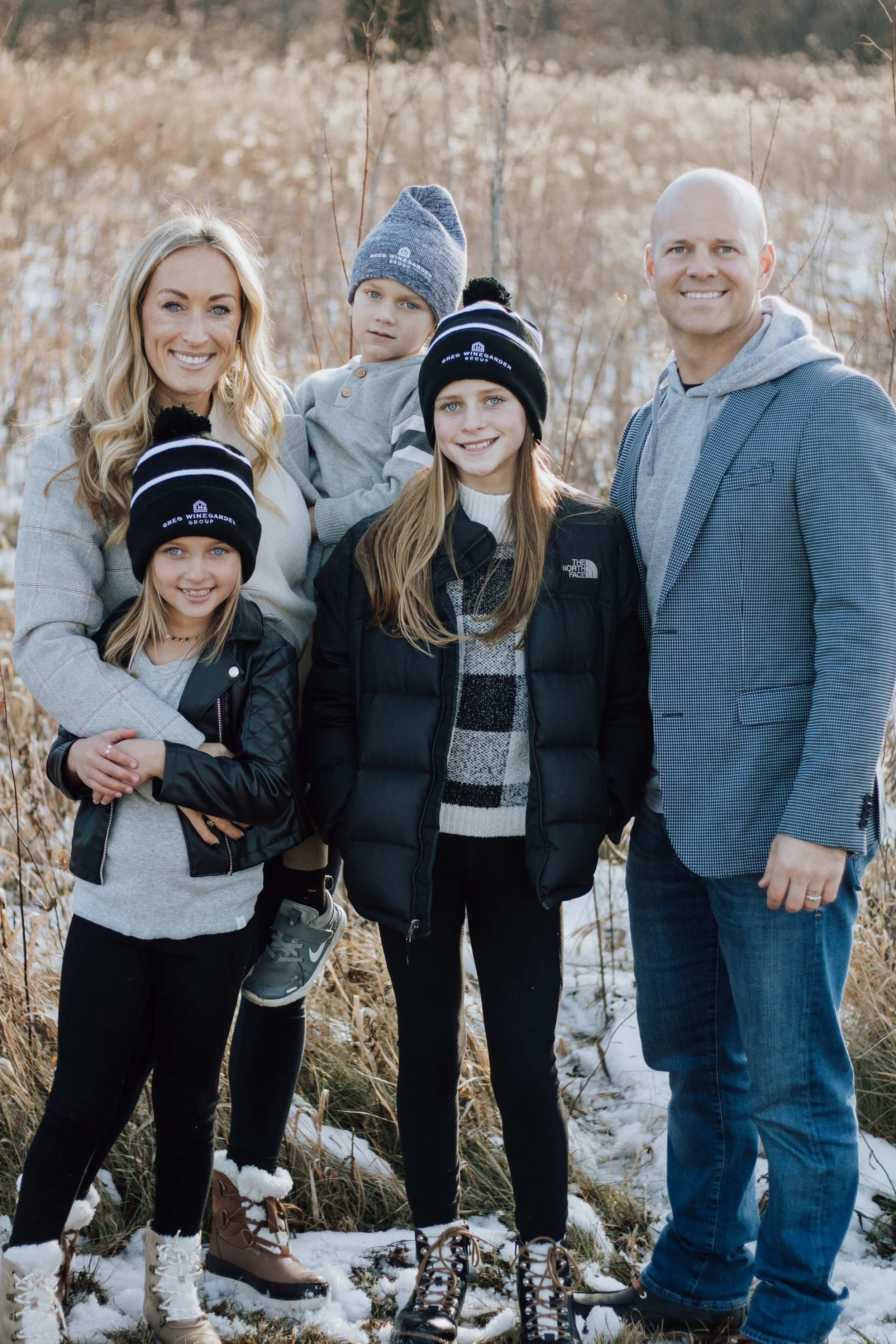 Family of six outdoors in winter, standing on snow-covered ground among tall grasses with bare trees in the background. The family includes a mother, father, and four children, all dressed in warm clothing, with three wearing black and white beanies.