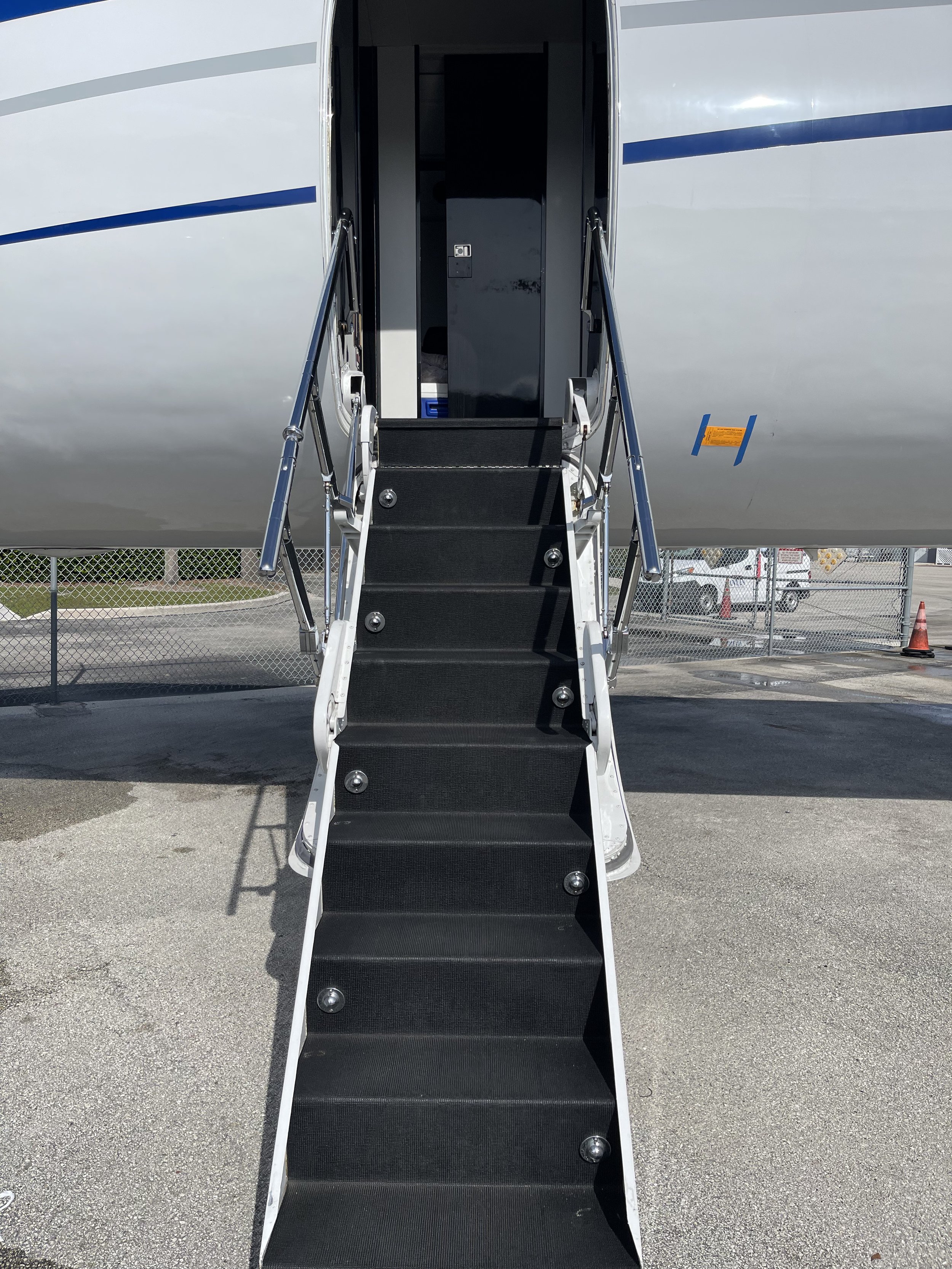 An airplane boarding staircase leading up to an aircraft door, with a fence and parking lot visible in the background.