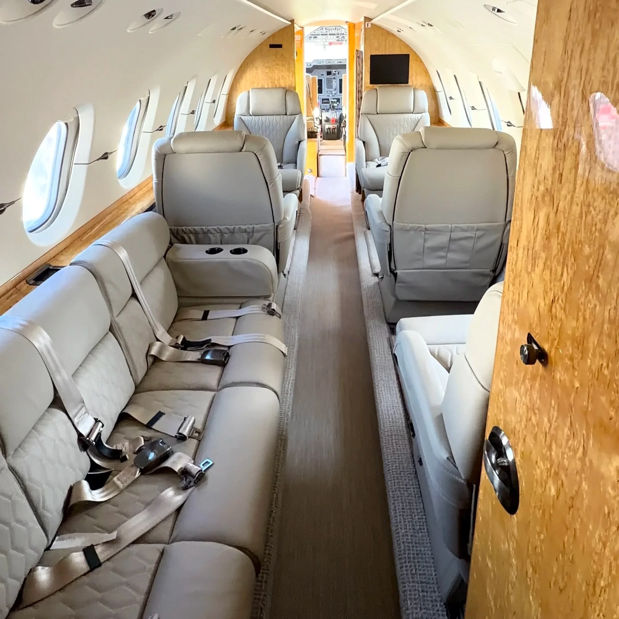 Inside the fuselage of an airplane, showing white leather seats with seat belts, oval windows on the left, and a cockpit visible in the background.