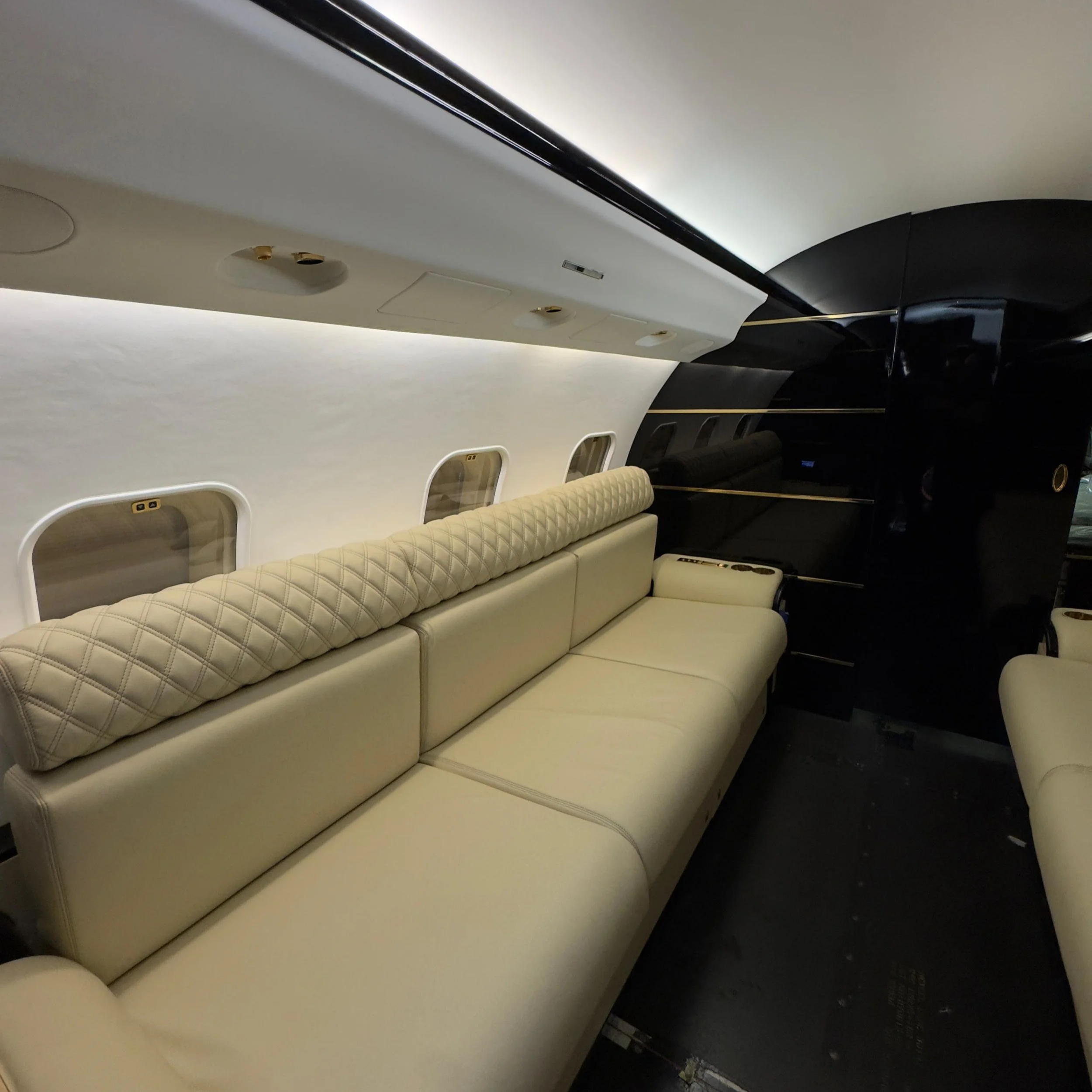 Interior of an airplane cabin showing a row of cream-colored quilted seats, with small over-head compartments, windows, and a dark ceiling section.
