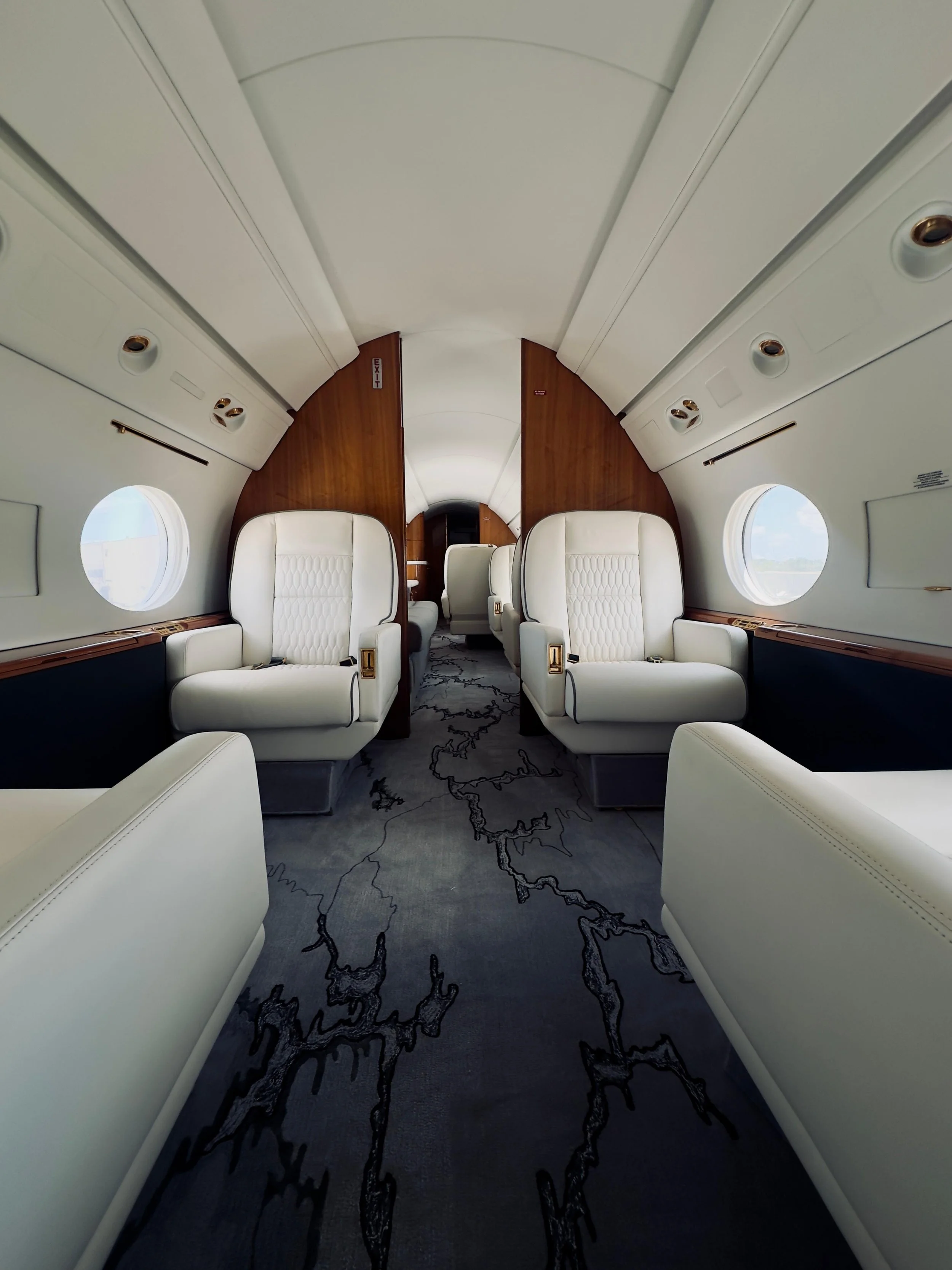 Interior of private jet with cream leather seats, wooden accents, round windows, and patterned carpet.