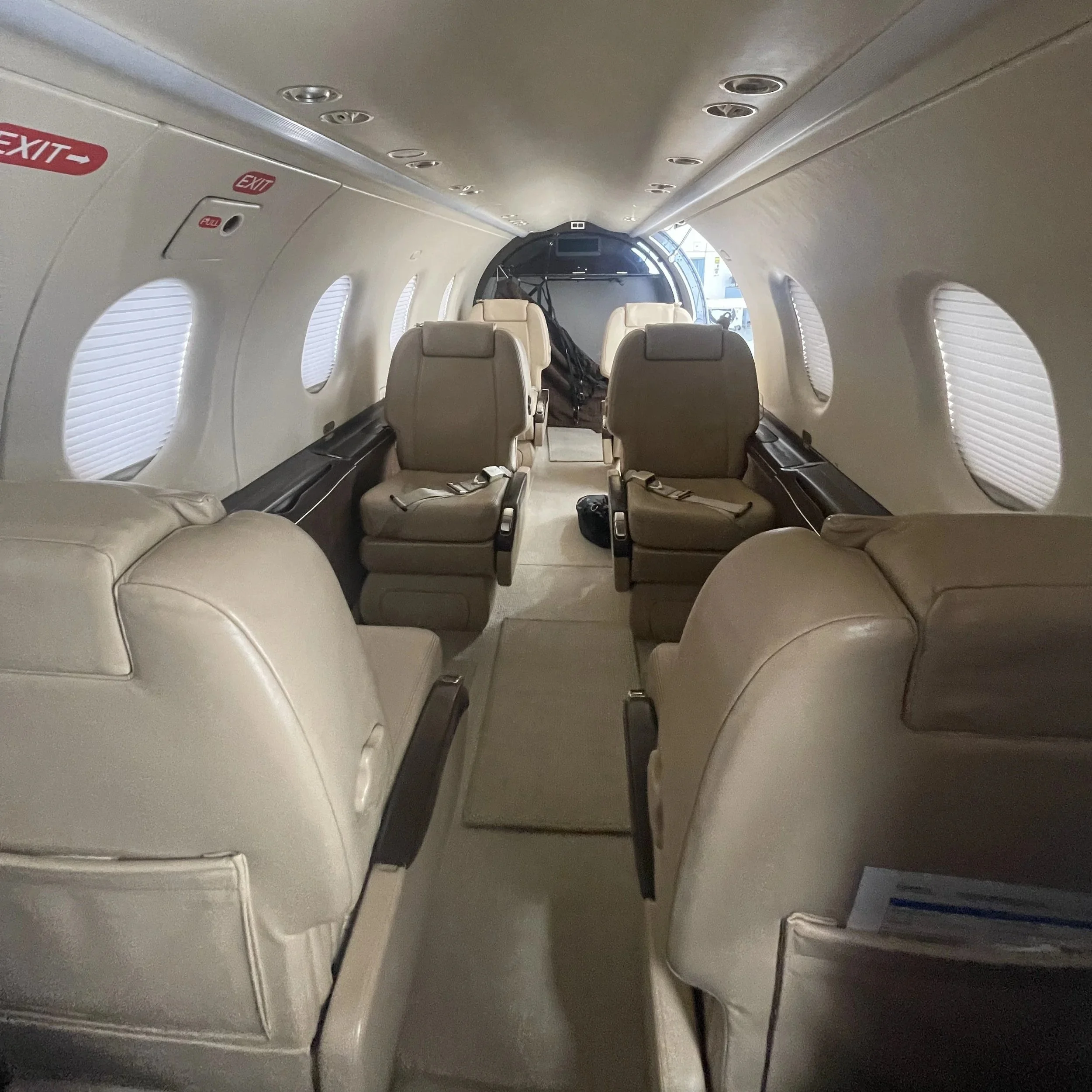 Interior of an airplane cabin showing several row of beige seats, side windows with blinds, overhead lights, and emergency exit signs.