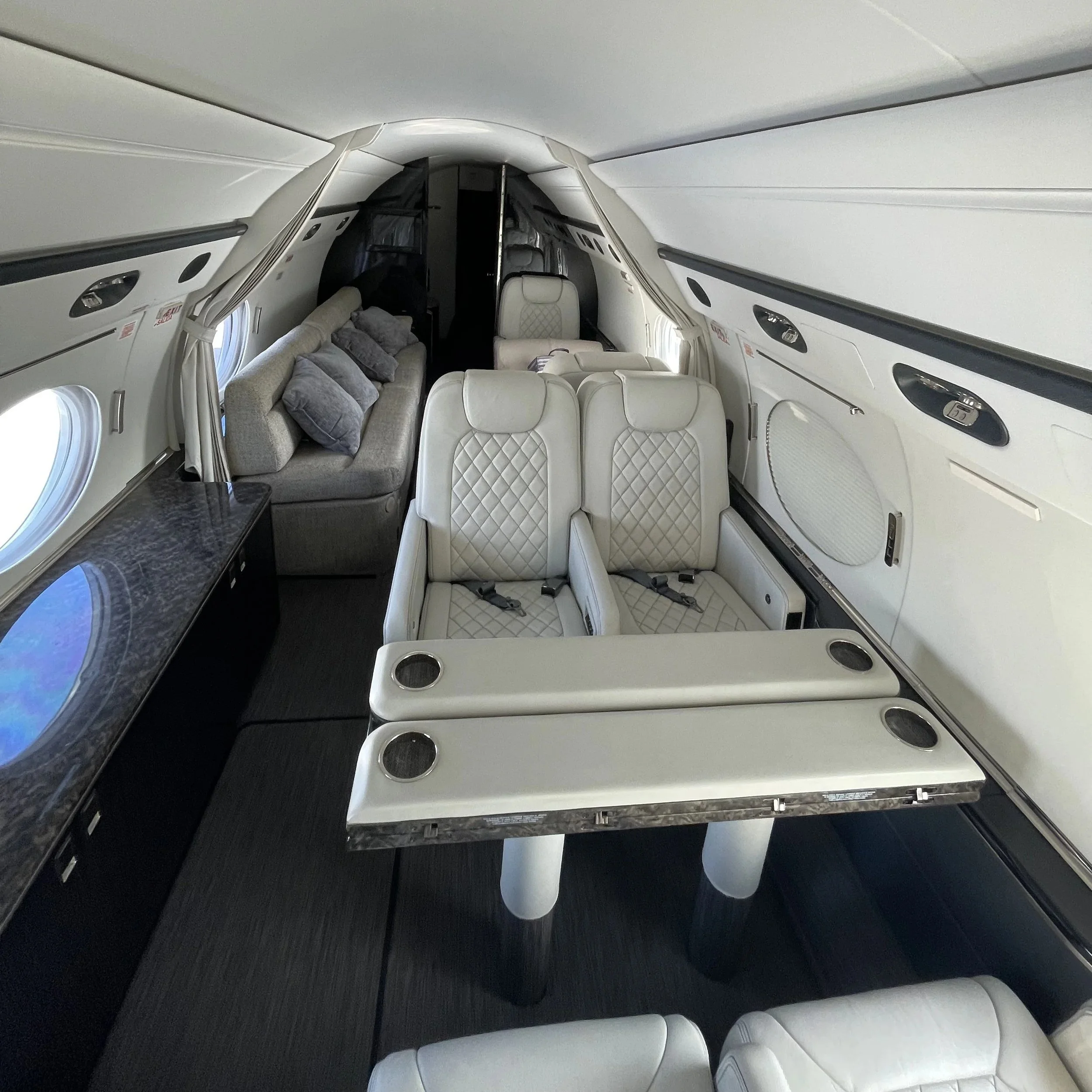 Interior of an airplane cabin with white leather seats, a gray sofa with pillows, and large windows on the sides.