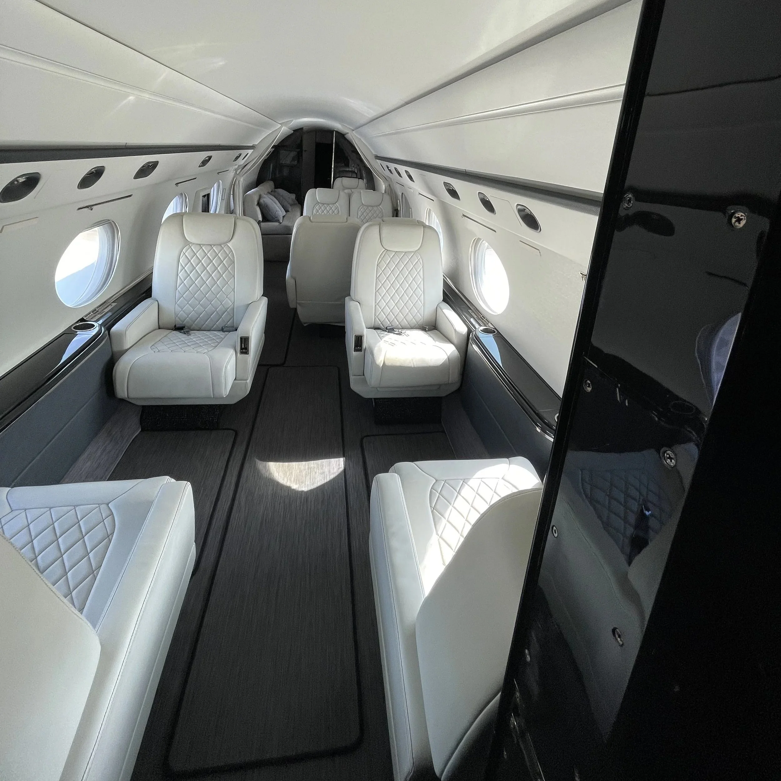 Interior of a private jet with white leather seats, large windows, and a large video screen on the right side.