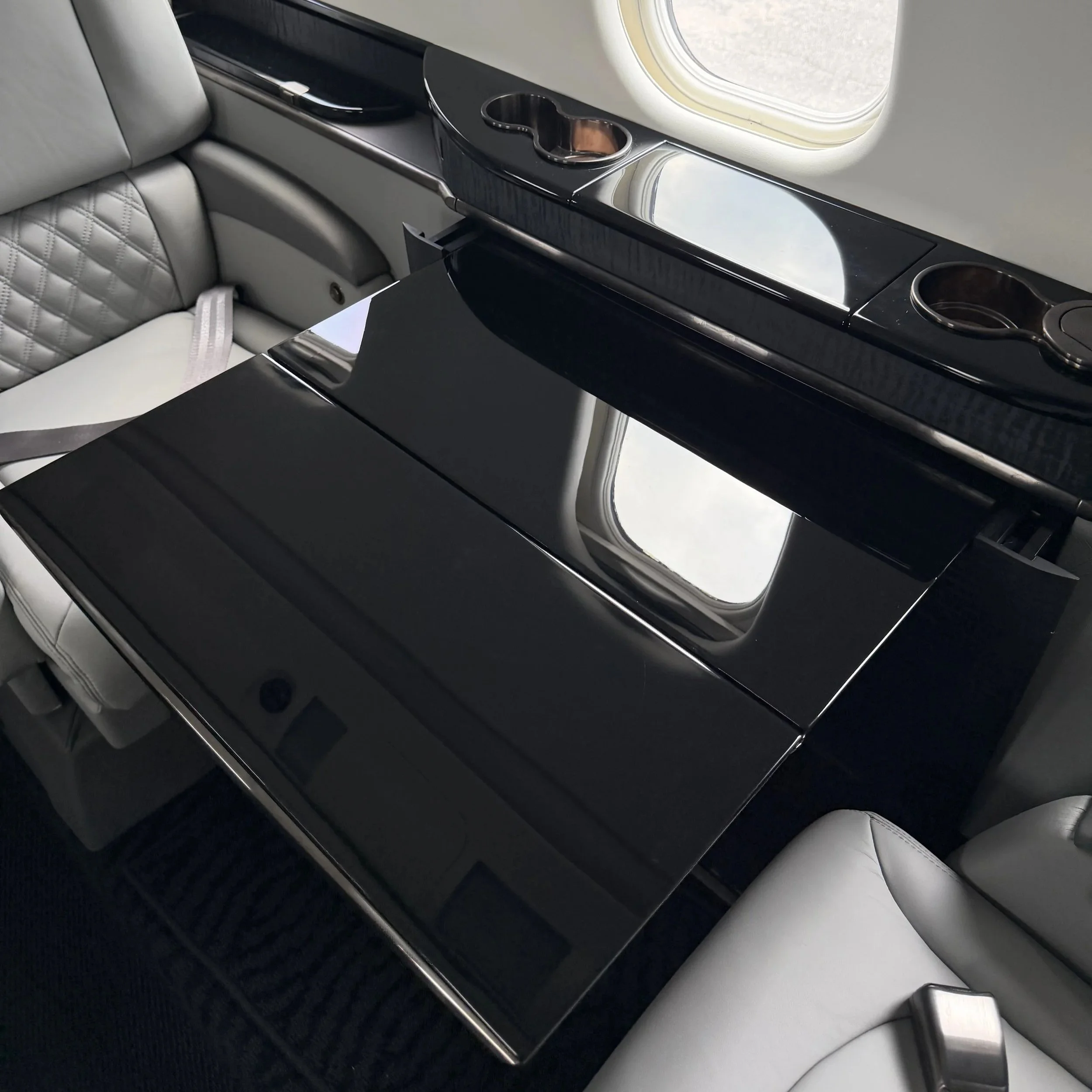 Inside the cabin of an airplane, showing a light gray leather seat with a quilted pattern on the backrest, a black glossy surface with built-in cup holders, and a window with a white frame.