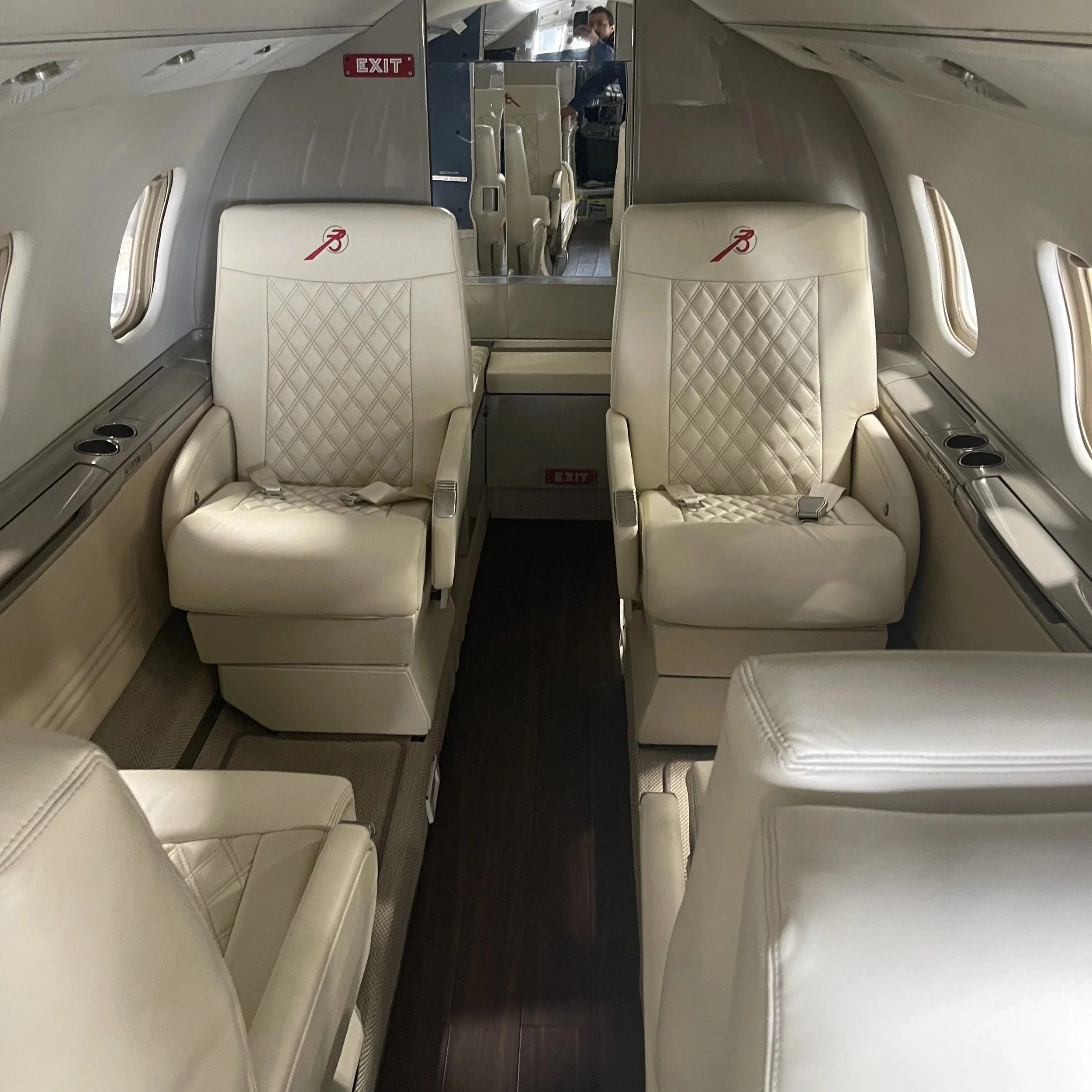 Interior of an airplane cabin with two beige leather seats in the foreground, small windows on both sides, and an emergency exit sign above a mirror reflecting a person taking the photo.