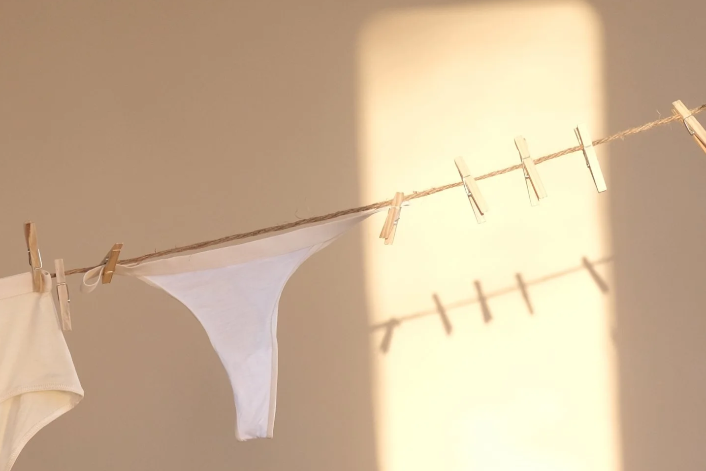 Clothesline with clothespins, holding white underwear, in front of a beige wall with sunlight and shadow.