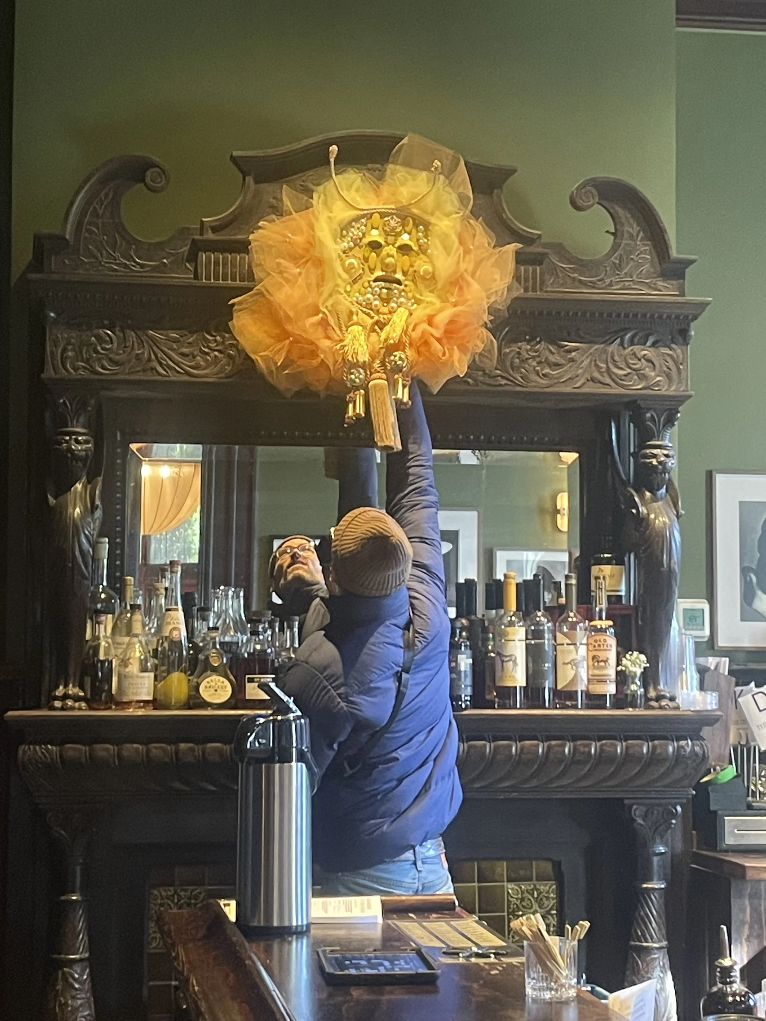 Installation in progress at The Chloe Hotel showing a sculptural mask being mounted above an ornate mirror behind the bar.