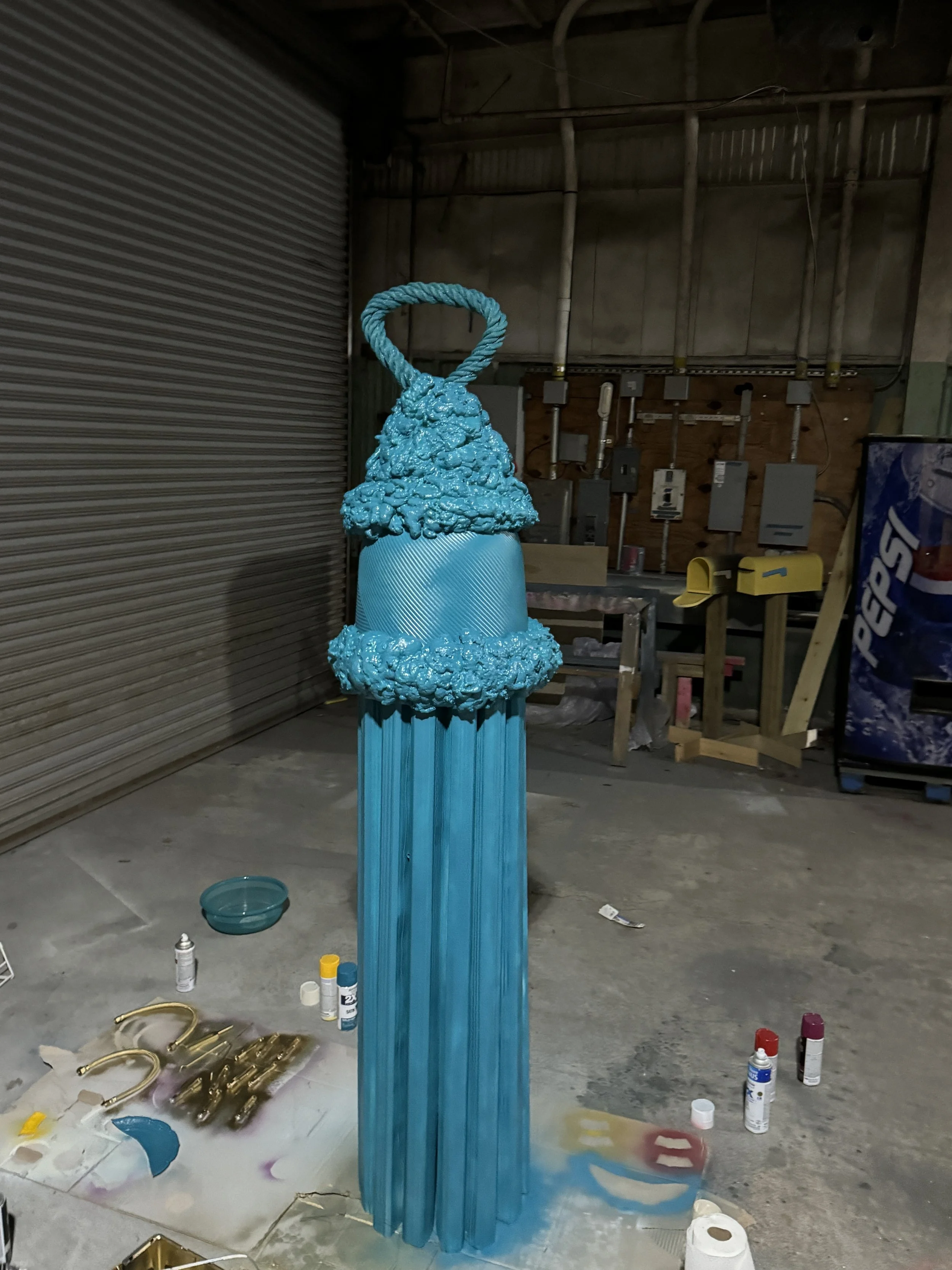 An over-sized tassel painted blue, made of found objects such as gutter siding, rope, and a flower pot.