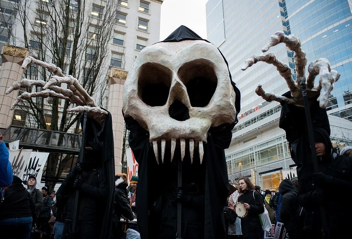 A giant skeleton puppet (grim reaper) operated by three people who are present at a political protest.