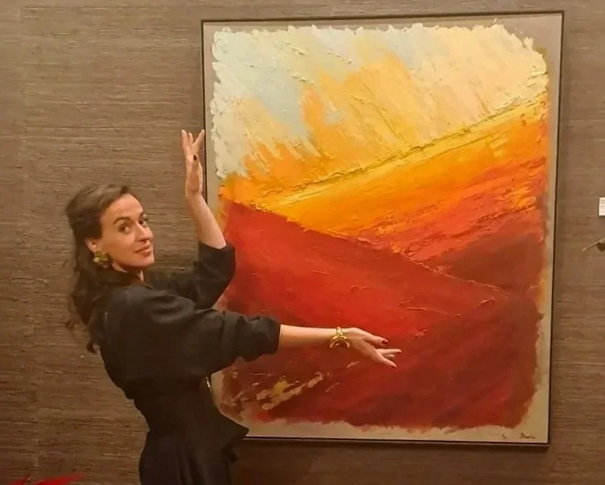 Una mujer posa junto a una pintura abstracta de colores cálidos, con tonos amarillos, anaranjados y rojos, en una galería de arte.