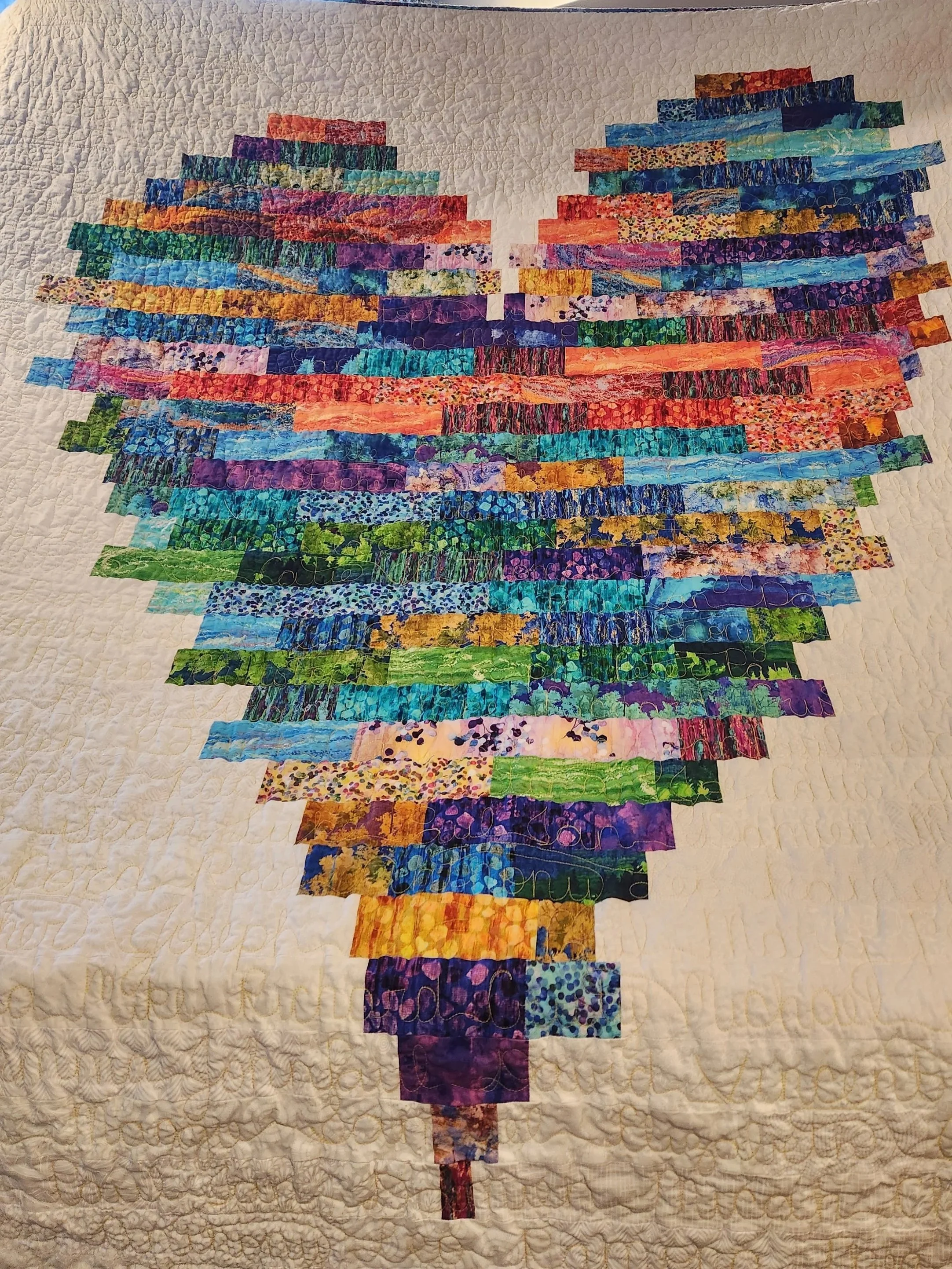 A quilt featuring a colorful, pixelated heart design made from various fabric strips on a beige background.