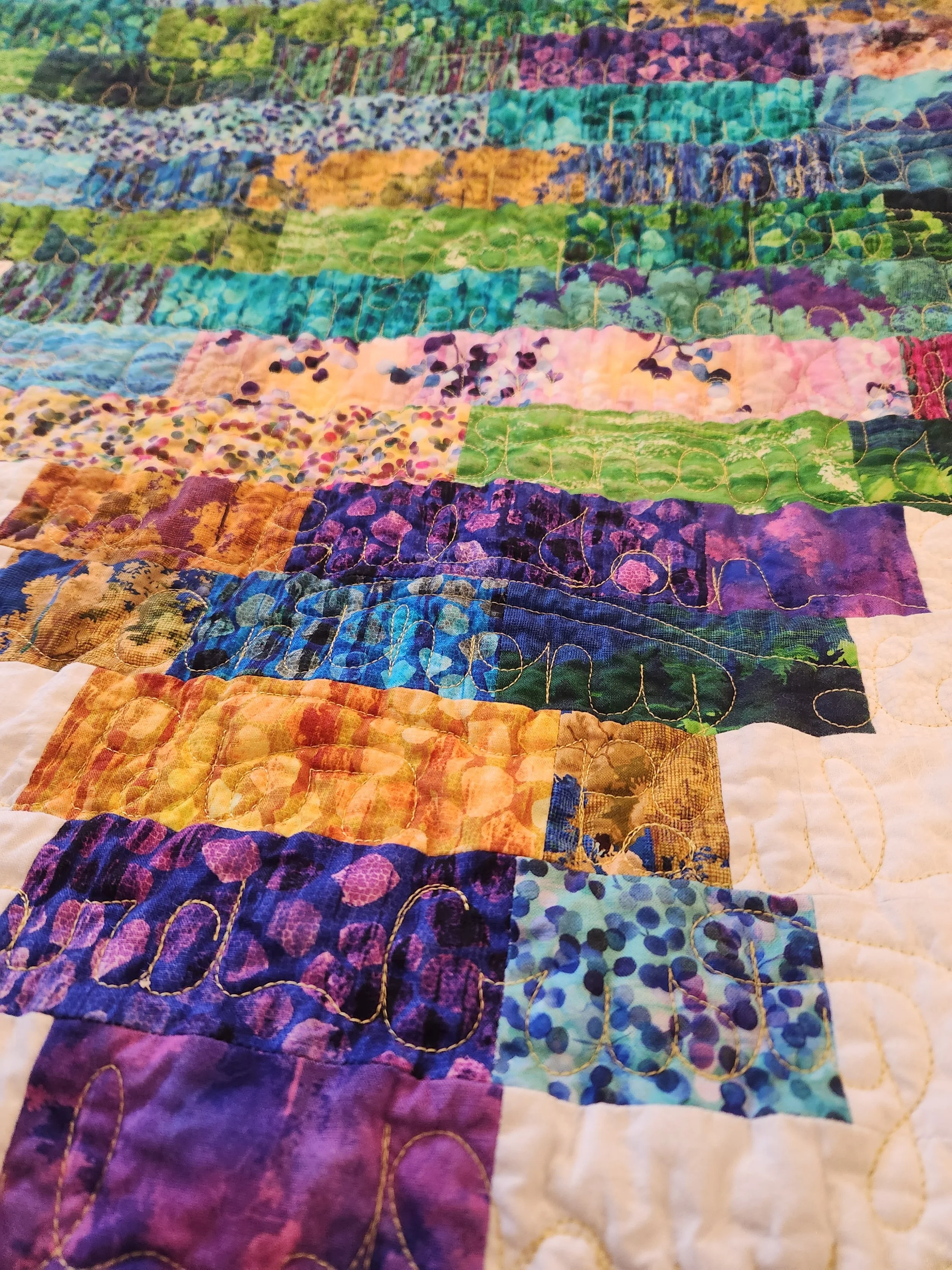 Close-up of a colorful quilt with various fabric patches in shades of blue, purple, green, pink, yellow, and orange, with intricate quilting stitches.