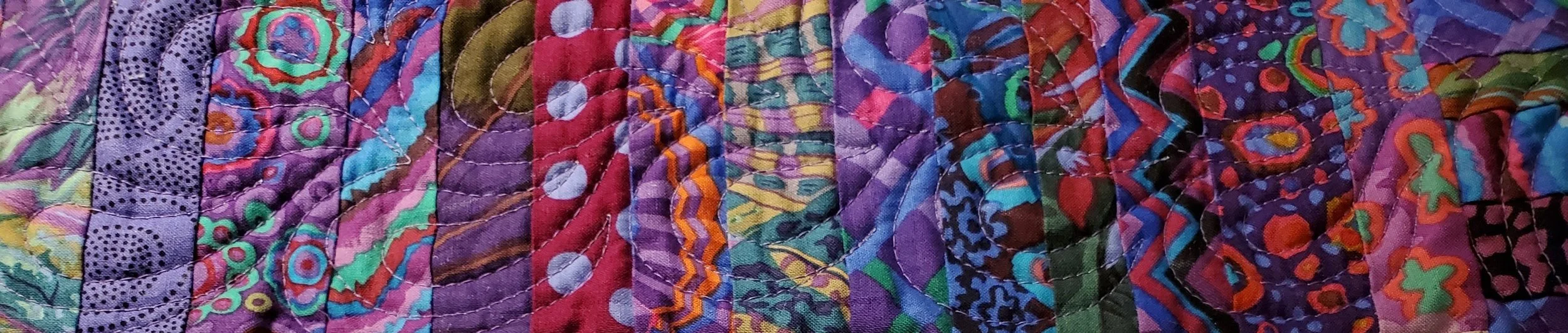 Close-up of a colorful quilt with intricate, abstract, and geometric patterns in various hues including purple, red, green, blue, and black.