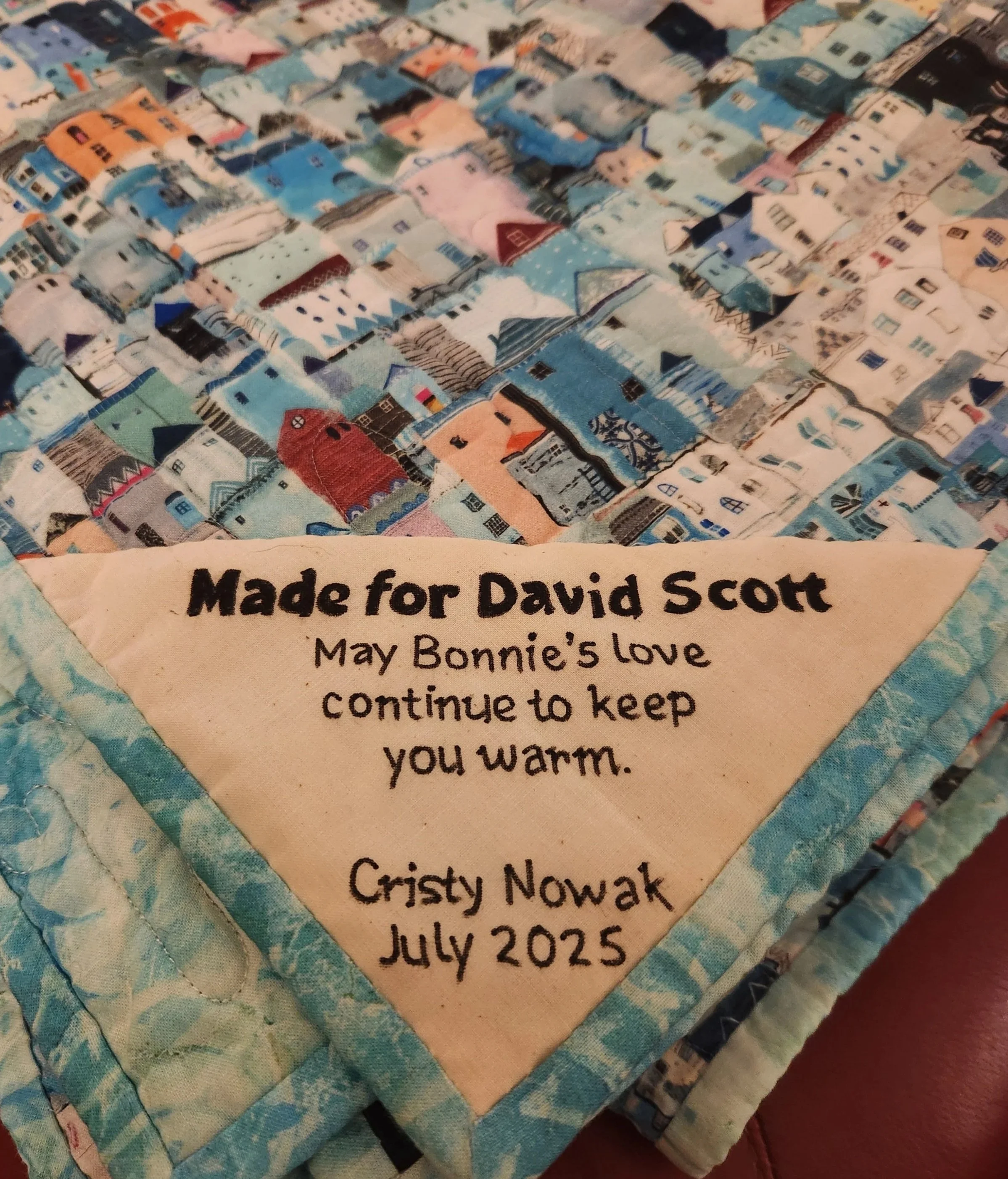 A quilt with colorful patchwork houses and a label that reads, 'May Bonnie's love continue to keep you warm. Cristy Nowak, July 2025.'