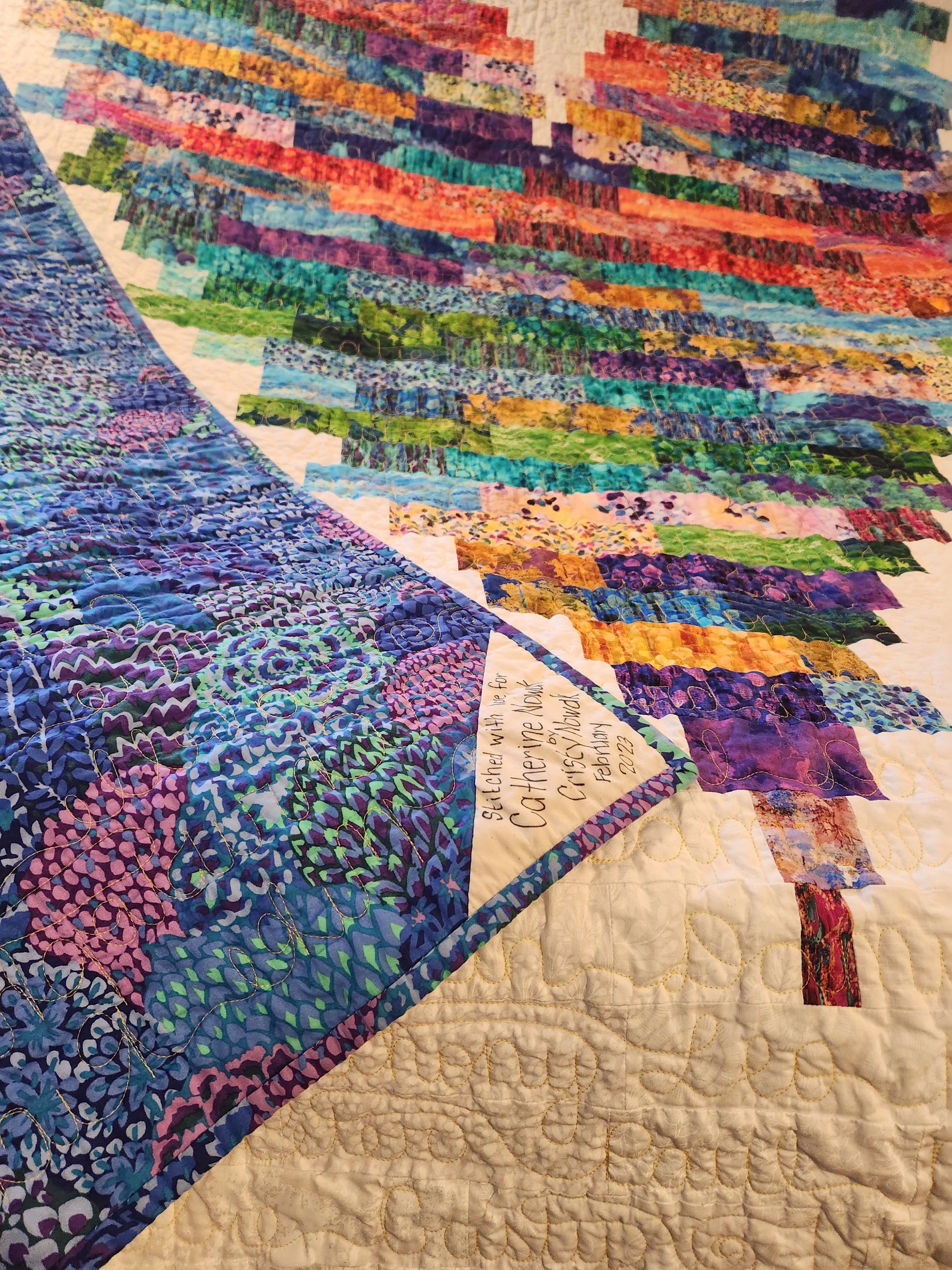 Close-up of a colorful quilt with various fabric patterns, including a section with an inscription reading 'Stitched with love for Catherine.'