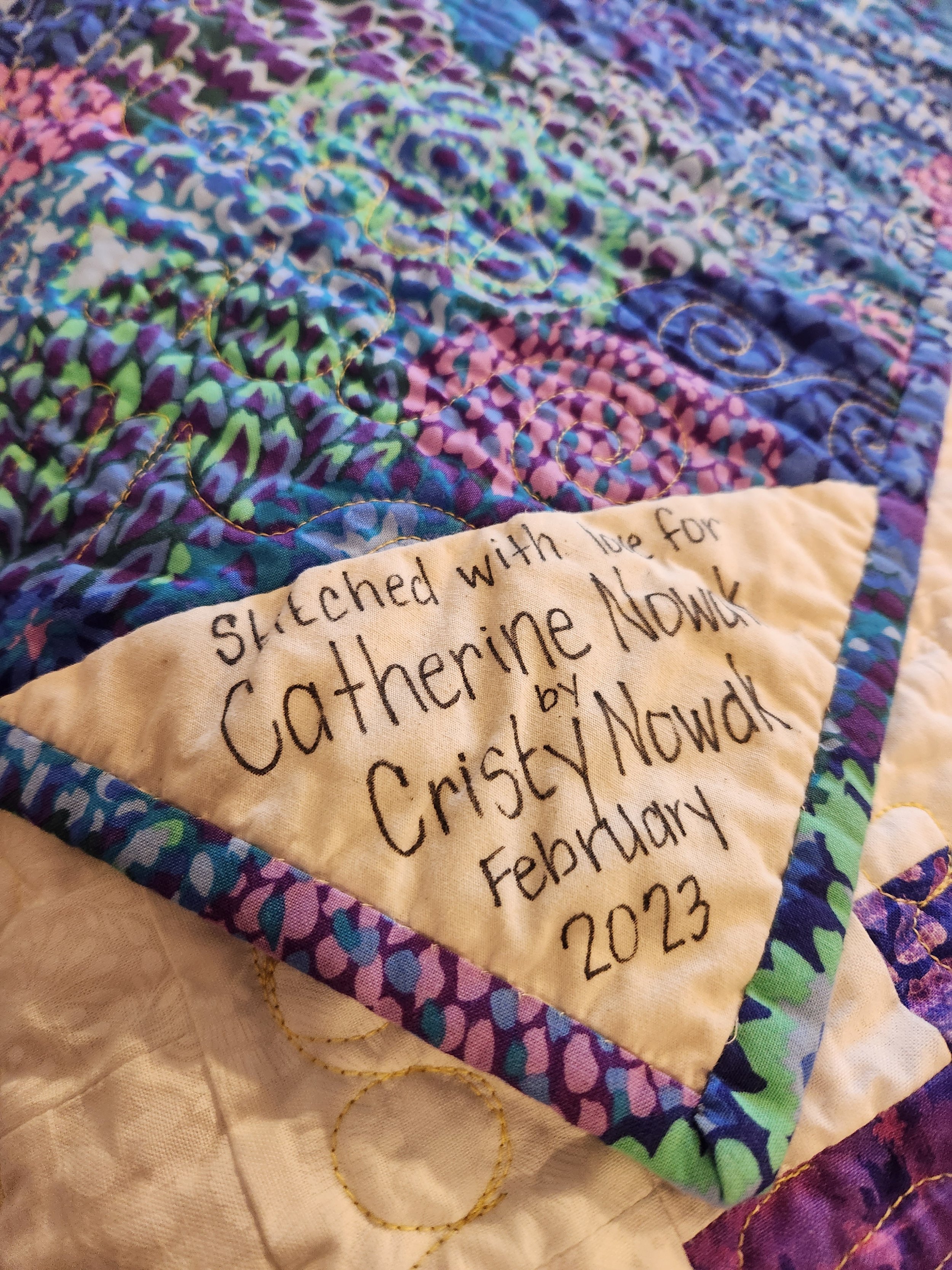A colorful quilt with a handwritten label that reads, 'stitched with love for Catherine Nowak by Cristy Nowak, February 2023.'