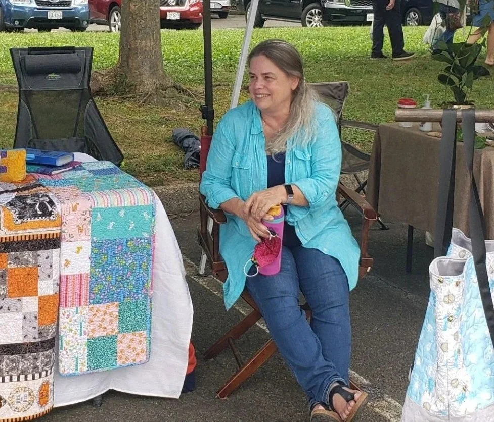 A woman with gray hair wearing a turquoise jacket sitting on a folding chair at an outdoor market, surrounded by tables with handmade quilts and crafts.
