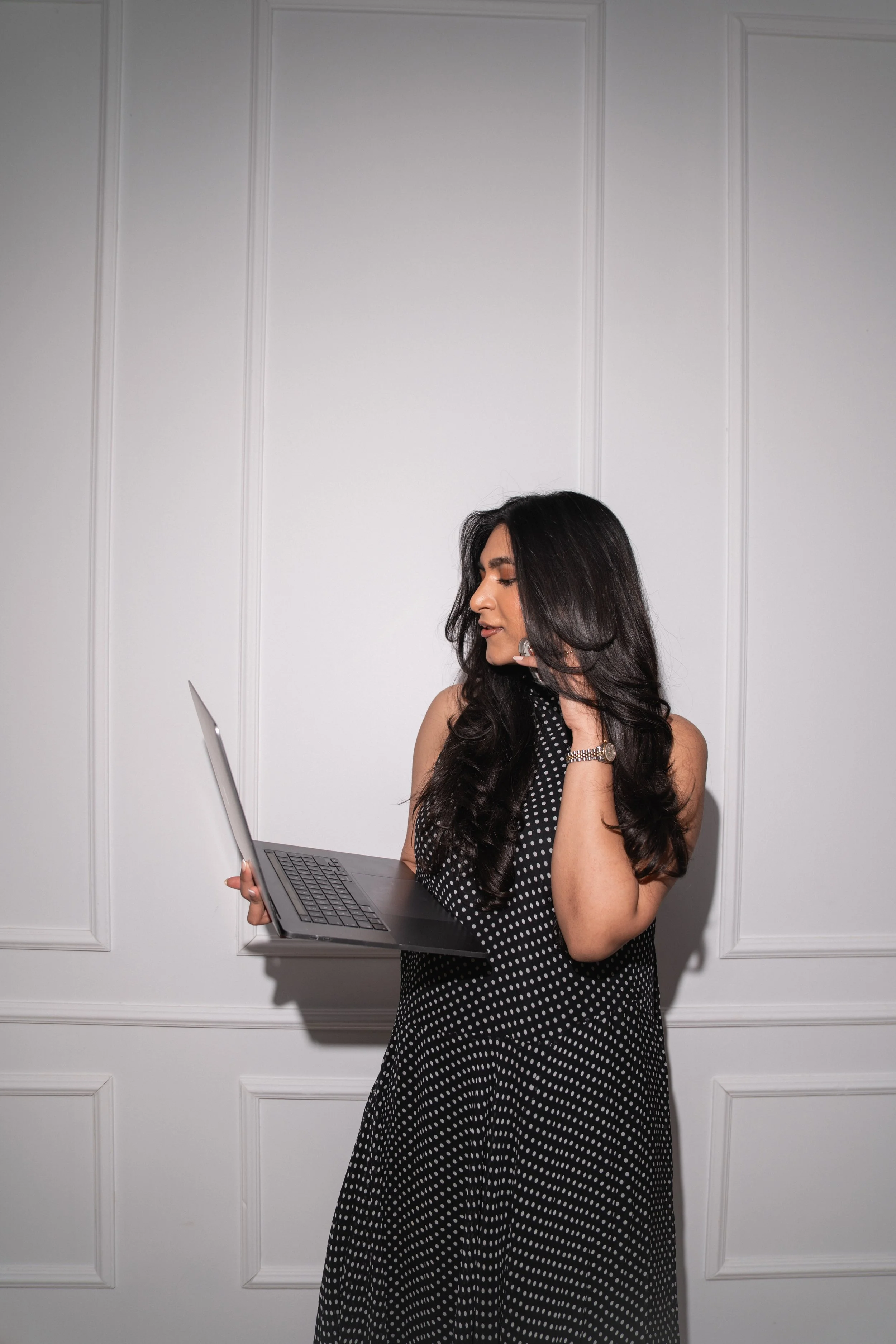 A woman with long dark curly hair wearing a black and white polka dot dress, holding a laptop and looking at it, standing in front of a white wall with decorative paneling.