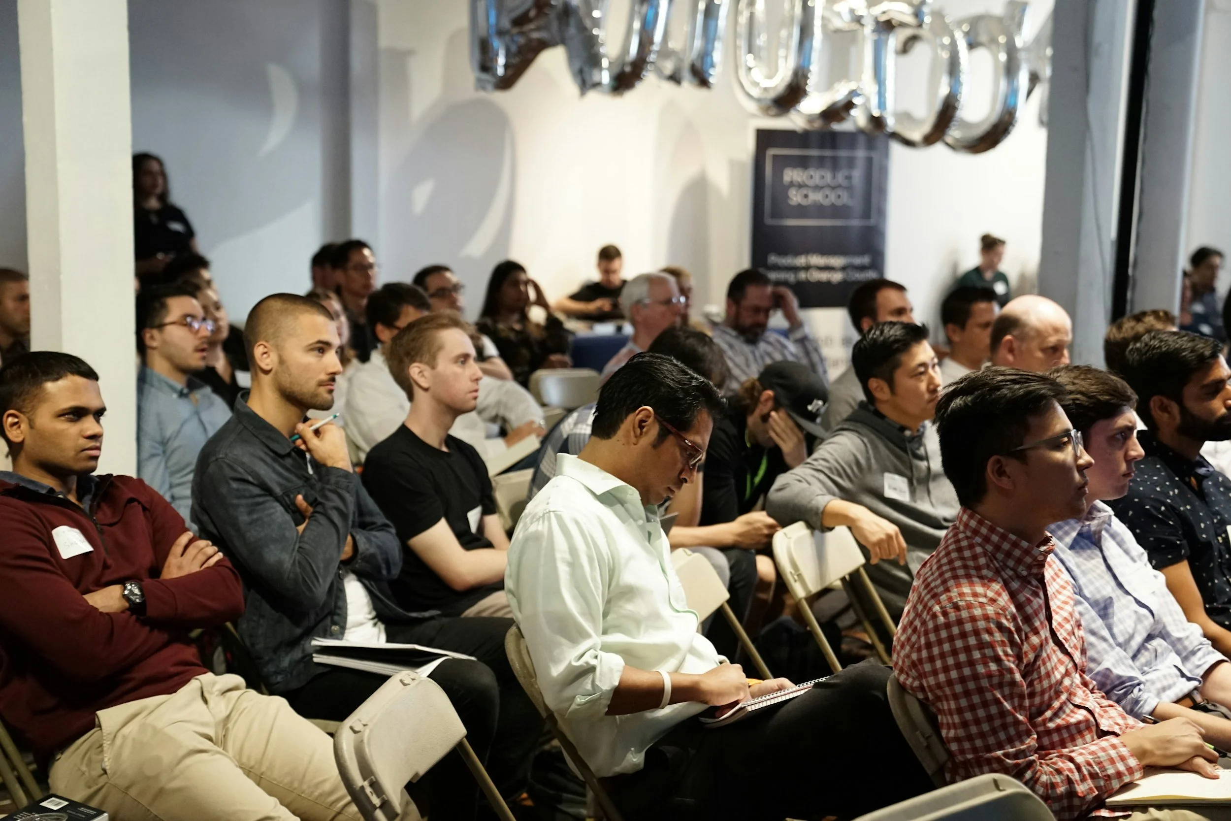 Audience attending a conference or seminar, seated in rows, some taking notes, in a room with white walls and balloons spelling out 'TALKS' at the front.