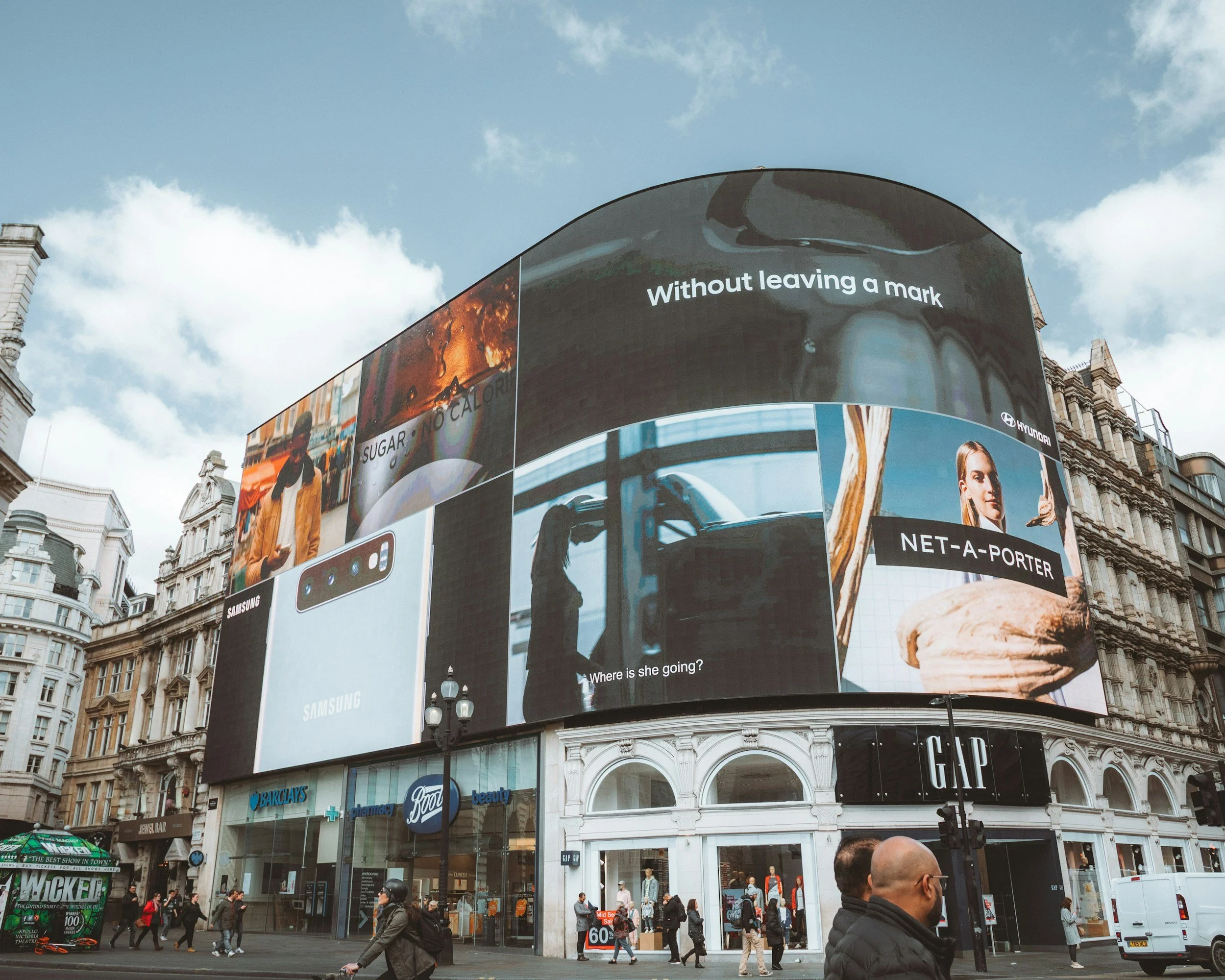 Large electronic billboard on a building in an urban area displaying multiple ads, including for Samsung, Hyundai, and Gap, with pedestrians and traffic below on a cloudy day.
