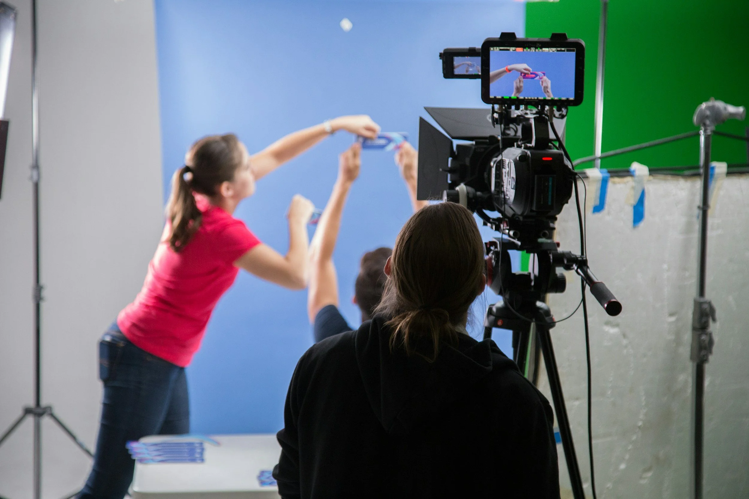 A woman in a pink shirt is taking a photo of a person with a tablet in front of a blue screen, while a camera records the scene behind her.