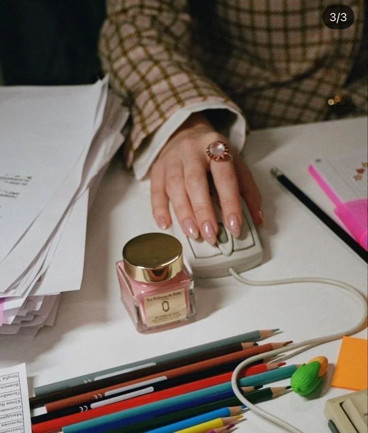 A person's hand with a large ring on the middle finger, typing on a computer mouse on a cluttered white desk with colored pencils, a pink nail polish jar, papers, and sticky notes.