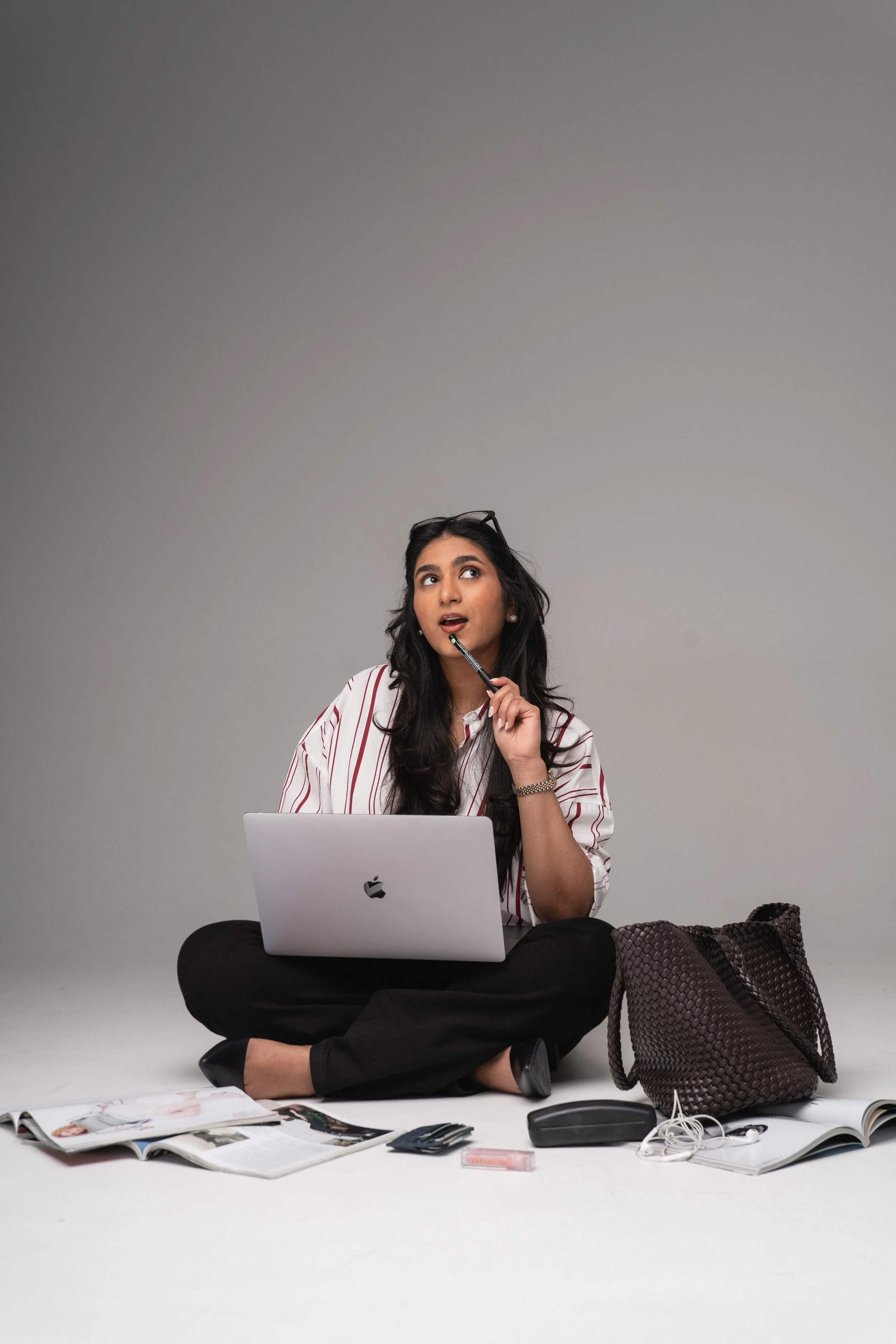 A young woman sitting cross-legged on the floor, using a laptop, surrounded by open magazines, a computer mouse, earbuds, and a woven bag, with a neutral gray background.