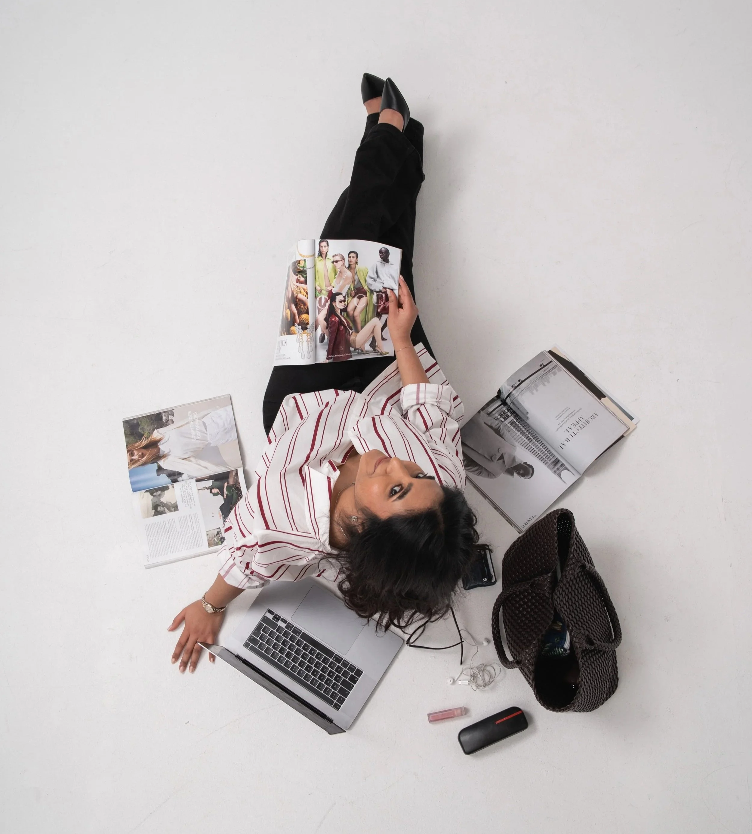 A woman lying on her back on a white surface, surrounded by magazines, a laptop, and personal items, with her head turned towards the camera. She is wearing a white shirt with red stripes and has long dark hair.