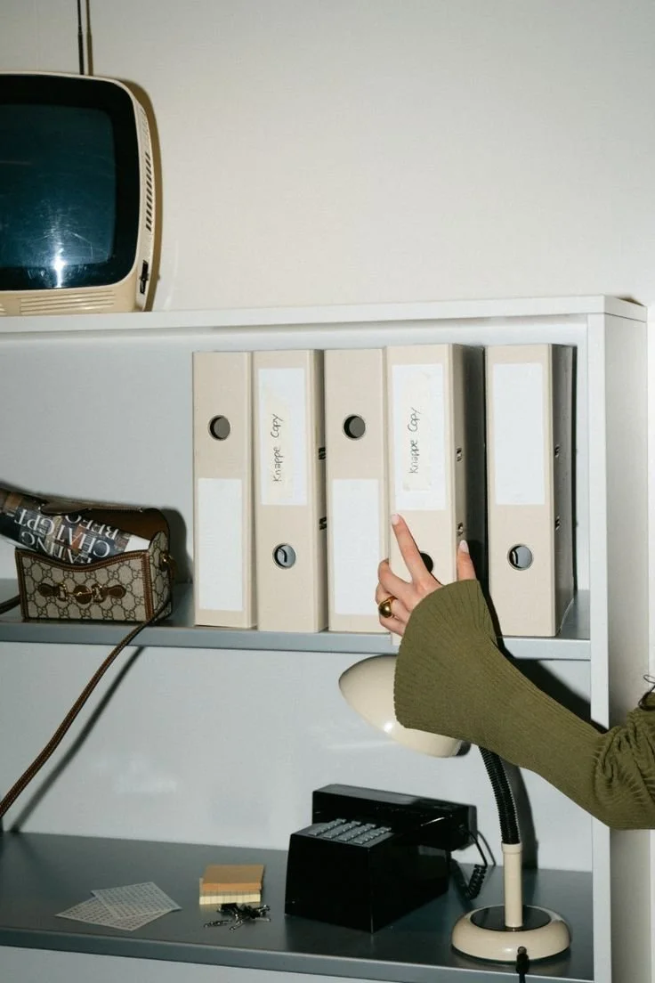 A person reaching for a white binder labeled "Knapek Copy" on a white office shelf, with a vintage TV on top, a designer handbag in a cubby, and office supplies on the desk below.