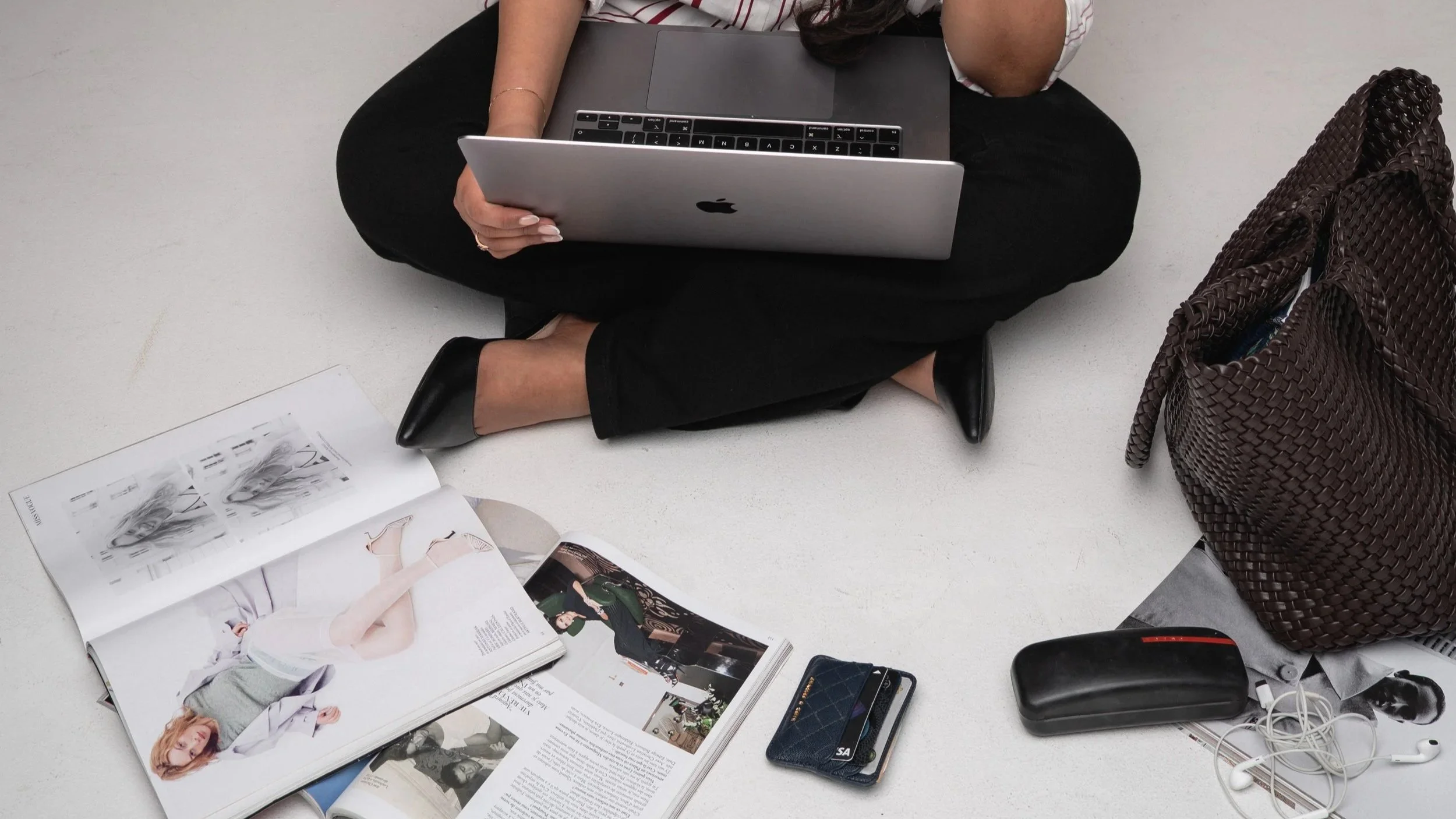A person sitting cross-legged on the floor using a laptop, with magazines, a purse, phone, earbuds, and other items scattered around them.