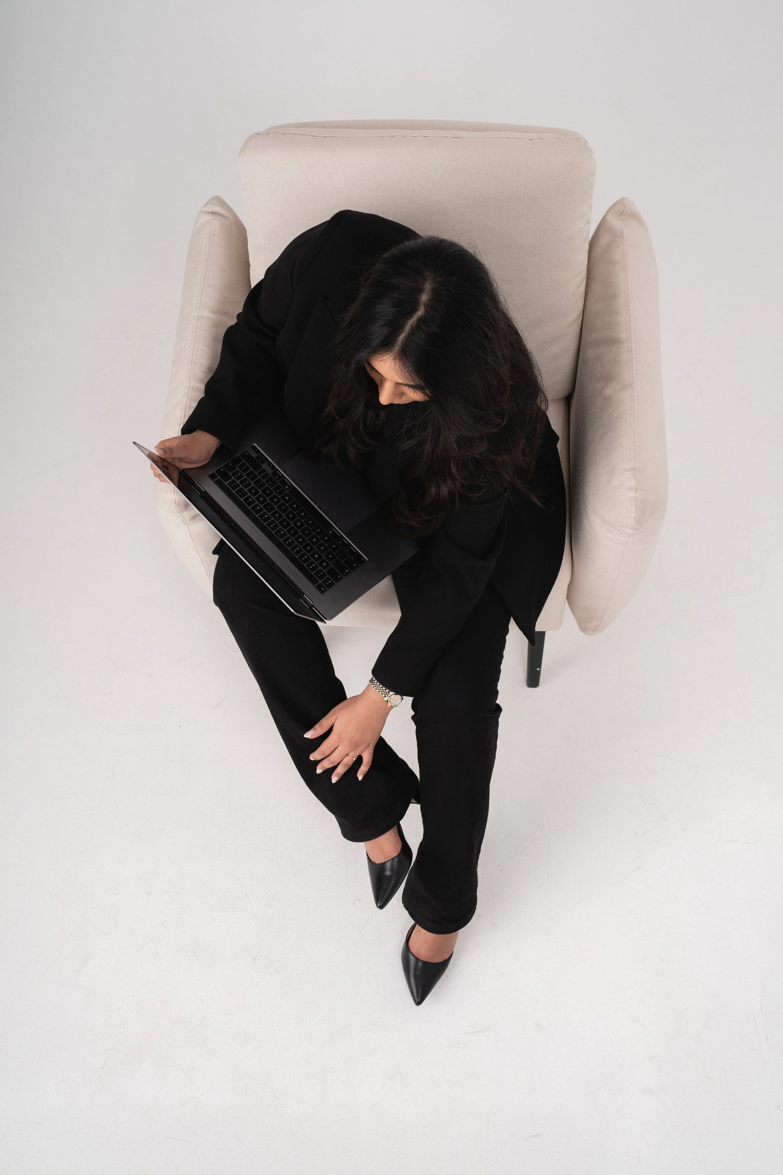 A woman in black clothing and black heels sitting in a light beige armchair with one leg crossed over the other and holding a laptop on her lap, viewed from above.