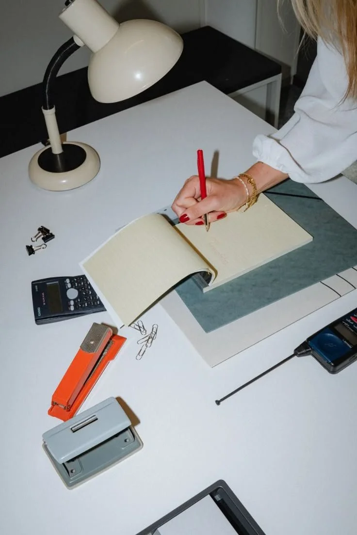 Woman writing in a notebook on a white desk, surrounded by office supplies including a lamp, calculator, paper clips, stapler, and a digital radio.