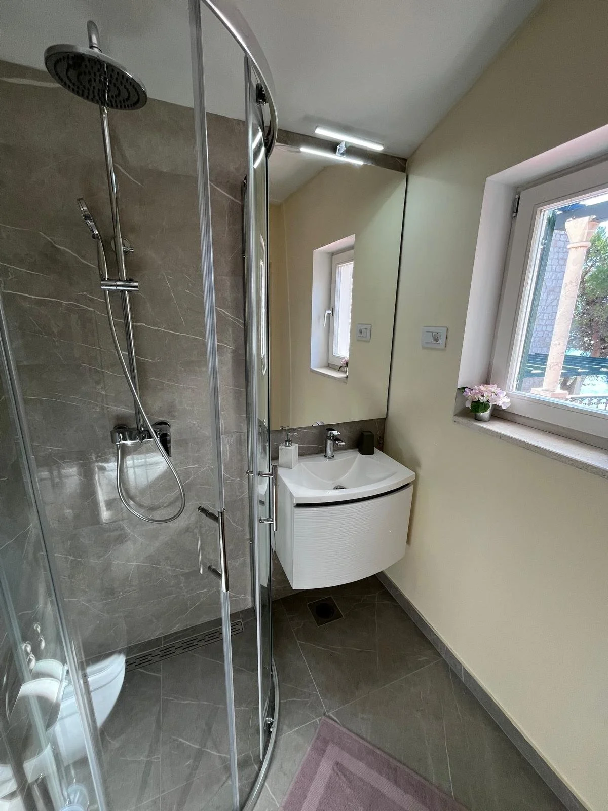 A small modern bathroom with a glass shower enclosure on the left, a white sink with a curved cabinet below in the center, a mirror above the sink, a small window with a flowerpot on the sill, and beige walls and floor tiles.