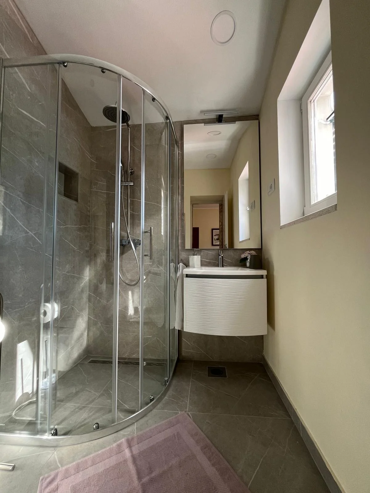 A bathroom with a curved glass shower enclosure, a vanity with a mirror, a window, and a pink rug on the floor.
