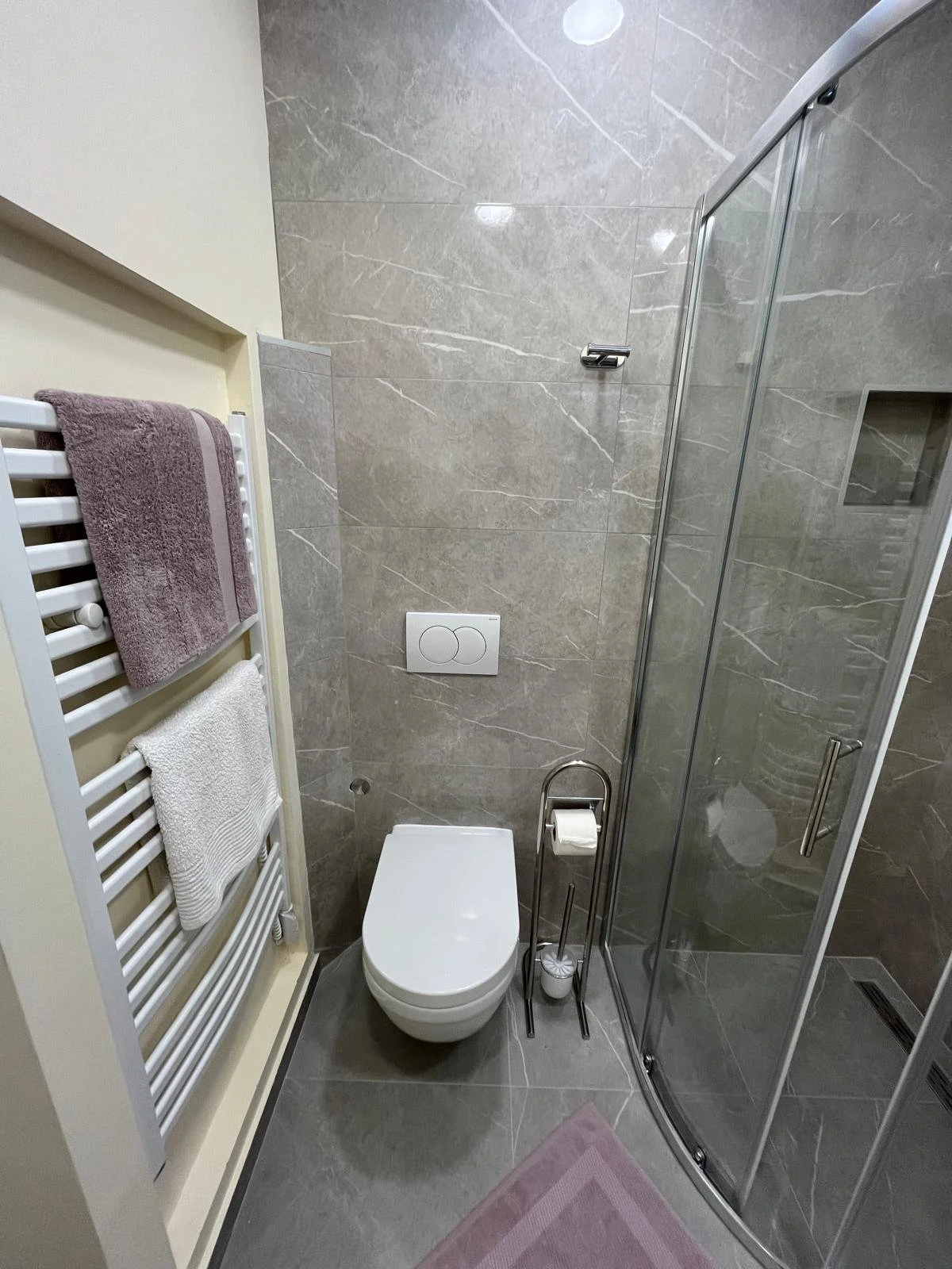 Small bathroom with gray marble wall and floor, white toilet, glass shower enclosure, white heated towel rail with towels, pink bath mat, toilet paper holder, and a small built-in shelf in the shower.