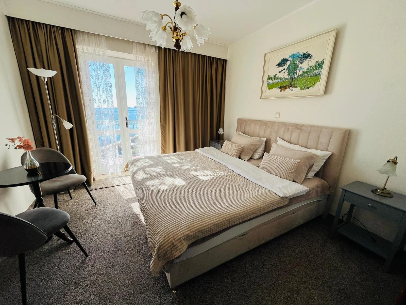 A hotel room with a large bed, beige headboard, white and beige bedding, and several pillows. To the side are two small gray nightstands with lamps. There is a window with sheer and heavy curtains, and a balcony door letting in sunlight. Opposite the bed, there is a small round table with two chairs. A framed landscape painting hangs above the bed. A chandelier hangs from the ceiling, and there is a floor lamp near the window.